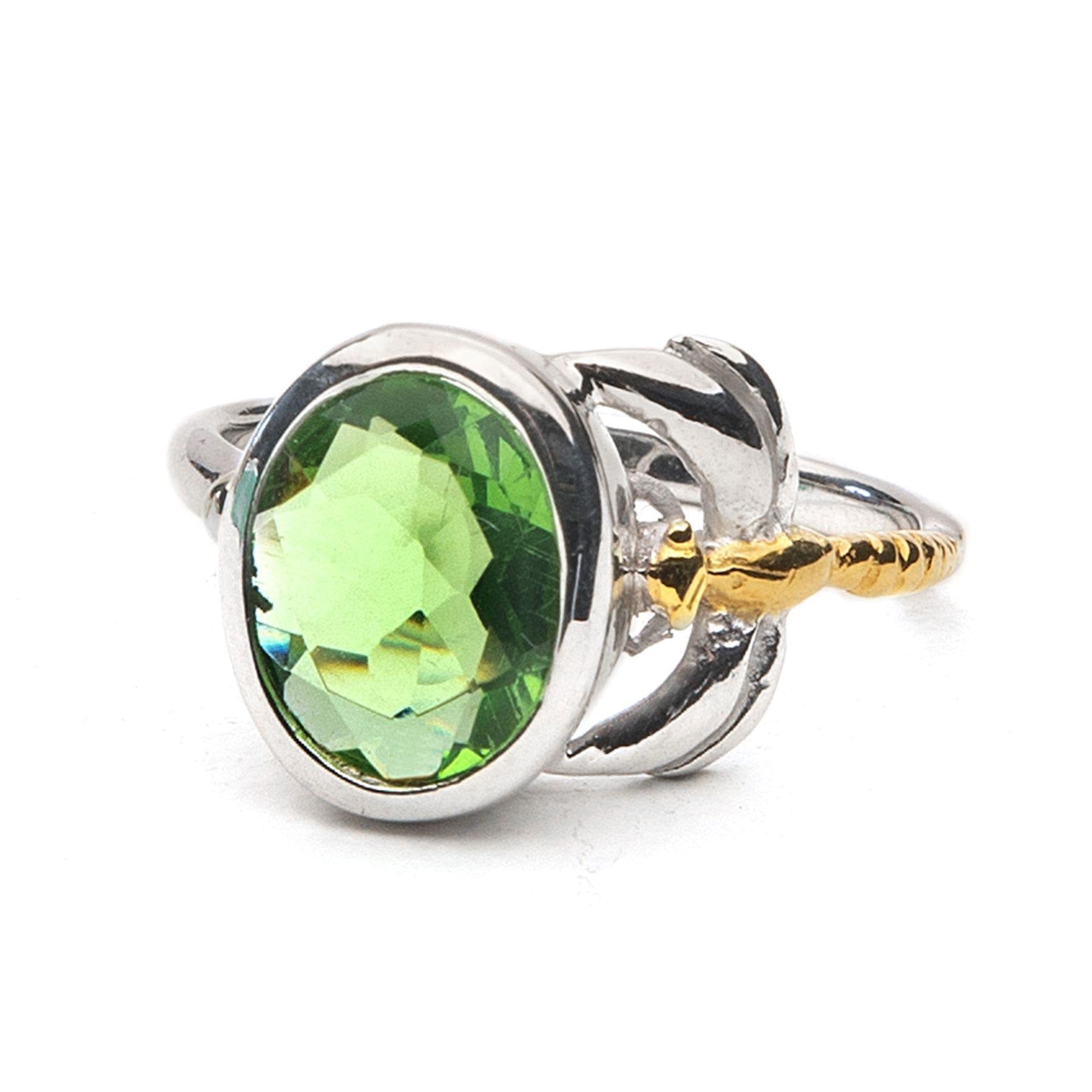 The Daydream Ring in various gemstones by Gallardo & Blaine Designs features a large, round, faceted green gemstone in the center. The setting has a unique design that includes gold accent details on the sides, resembling small leaves or petals. The smoot