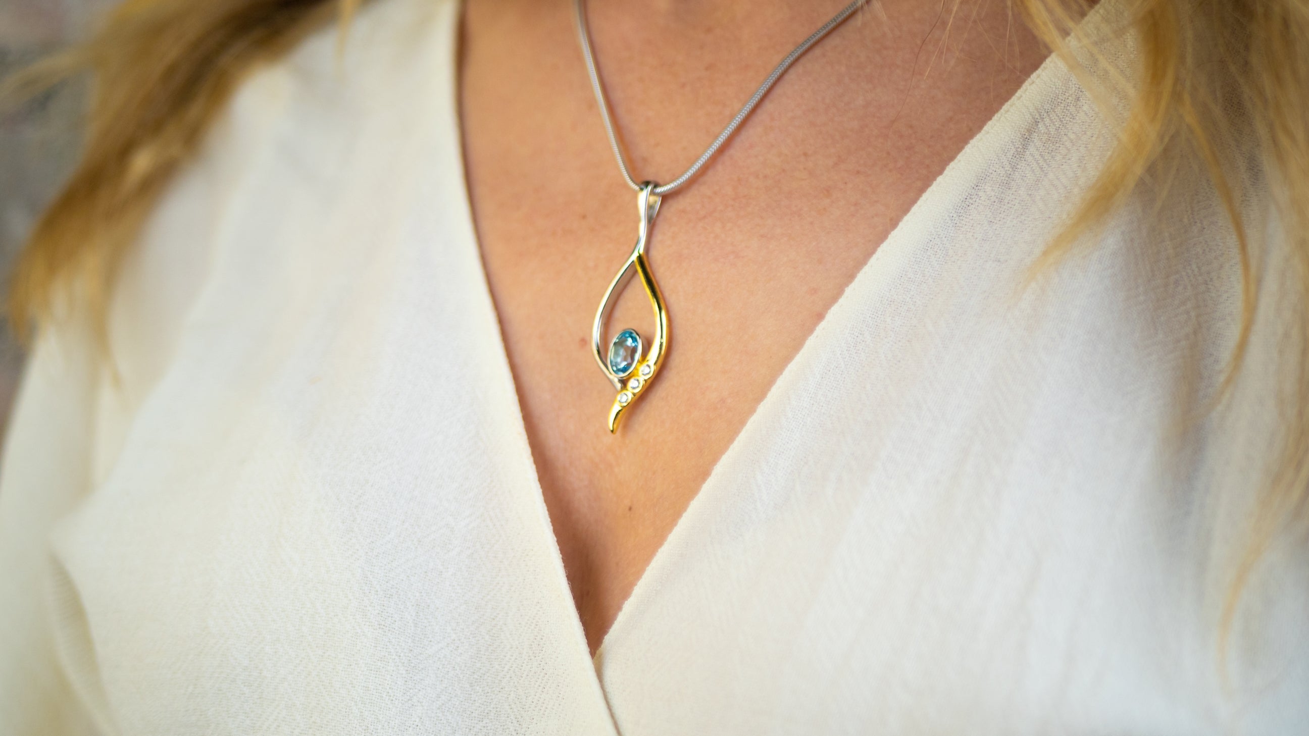 Close-up of a person wearing a sterling silver necklace with a teardrop-shaped pendant. The pendant, known as the Desert Star Pendant in various gemstones by Gallardo & Blaine Designs, features a small blue gemstone at the center and is worn over a white