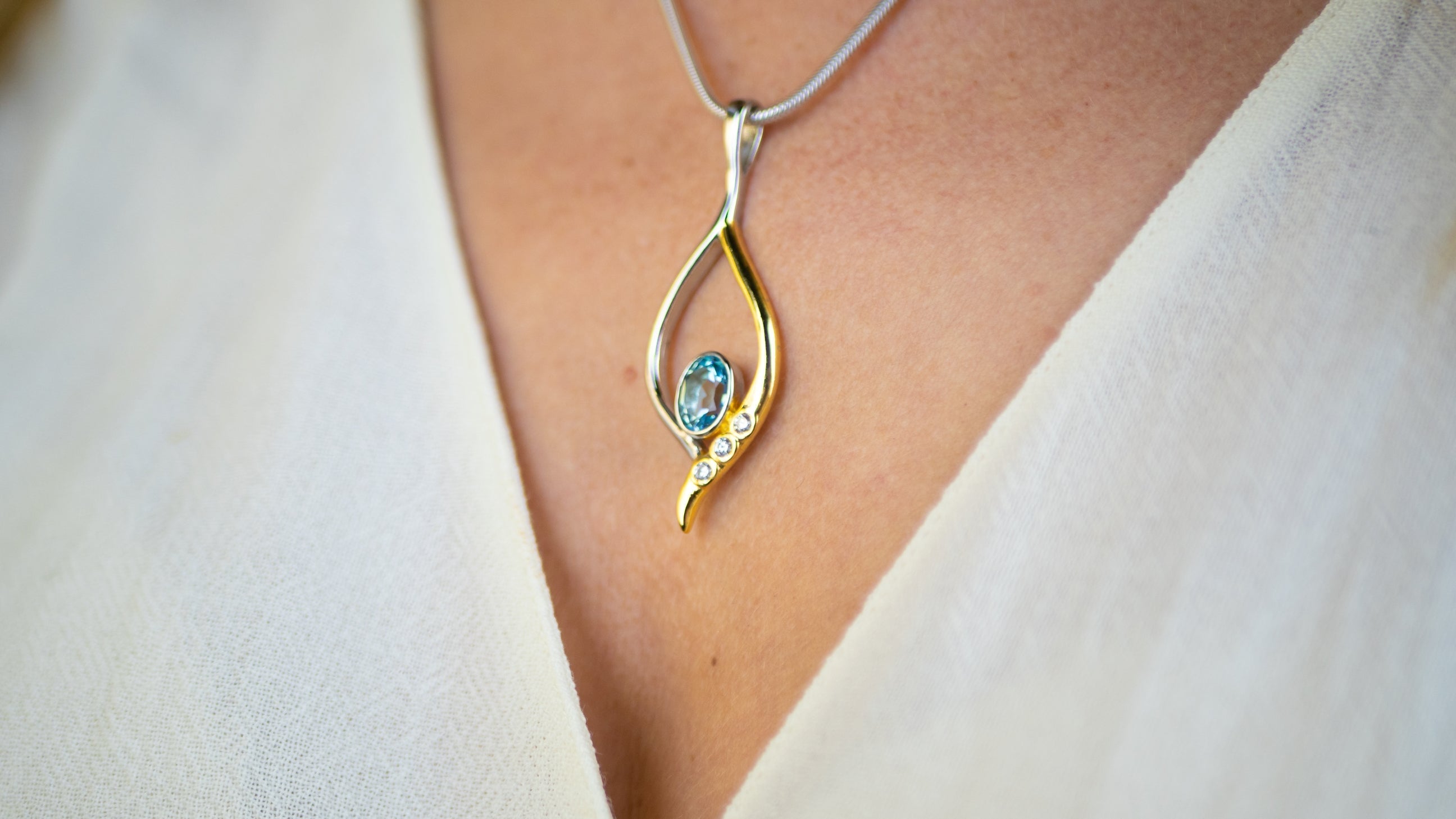 A close-up image of a person wearing a white garment, showcasing a delicate Desert Star Pendant in various gemstones by Gallardo & Blaine Designs. The pendant, crafted in gold vermeil and sterling silver, features a small blue gemstone at its center, ador