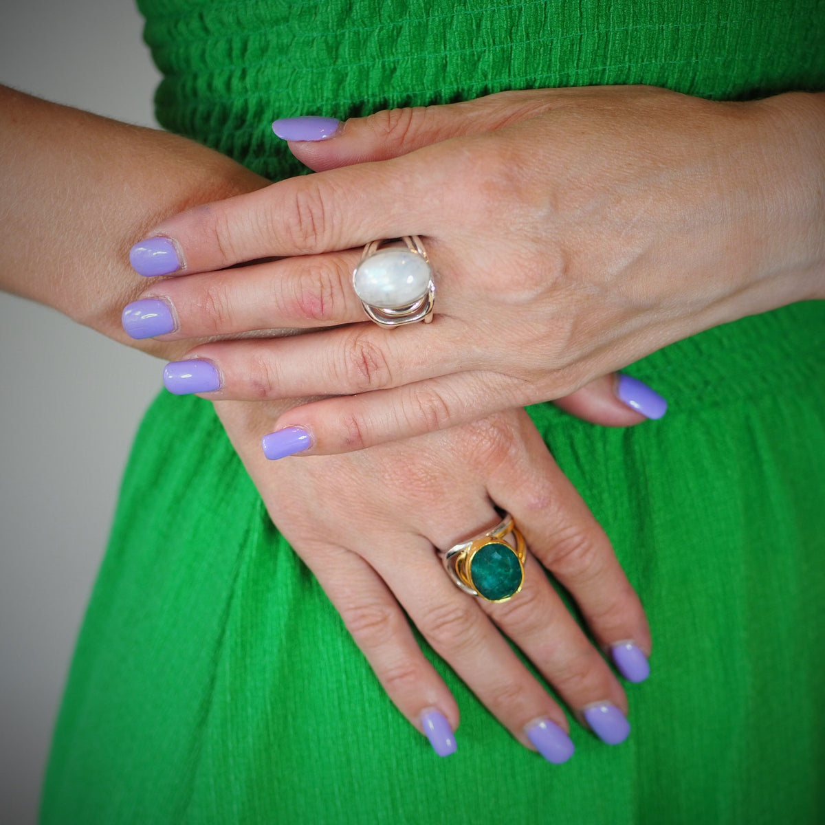 A person with light skin wearing a green dress has their hands crossed. They have lavender nail polish and are wearing two chunky statement rings: one Dewberry Ring in various gemstones by Gallardo & Blaine Designs with a large white stone on the middle f