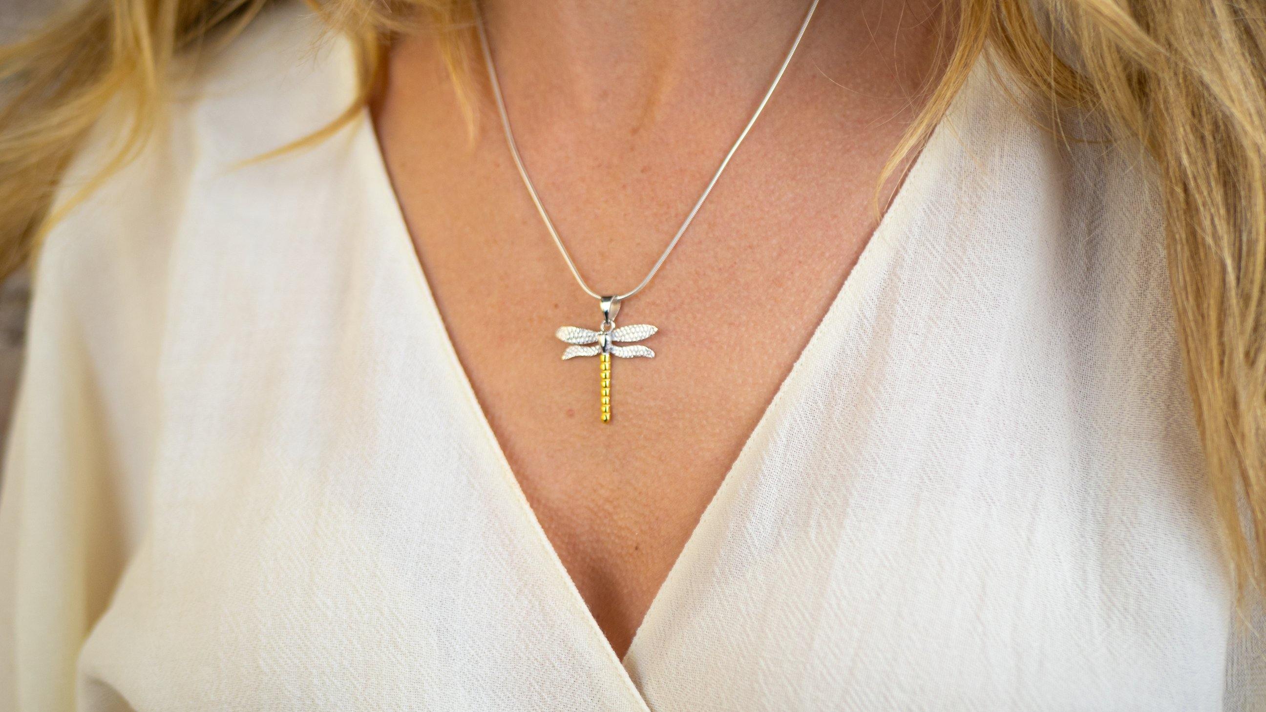 Close-up of a woman's neck adorned with a delicate silver snake chain featuring the Wildlife Pendant - Large Dragonfly With Chain from Gallardo & Blaine Designs. The dragonfly has detailed wings and a gold-toned body. She is dressed in a light beige, V-ne
