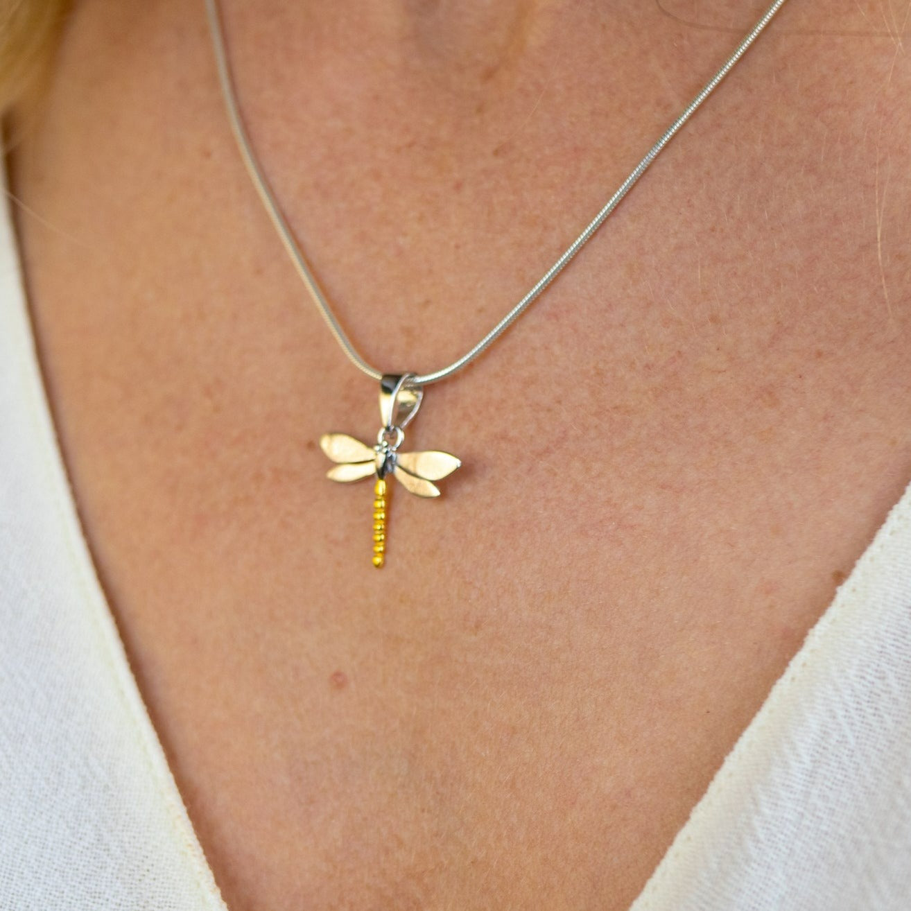 Close-up of a necklace from the Wildlife Pendant - Small Dragonfly With Chain by Gallardo & Blaine Designs, featuring a dragonfly pendant made of sterling silver and gold accents, worn by a person dressed in a light beige top. The background is softly blu