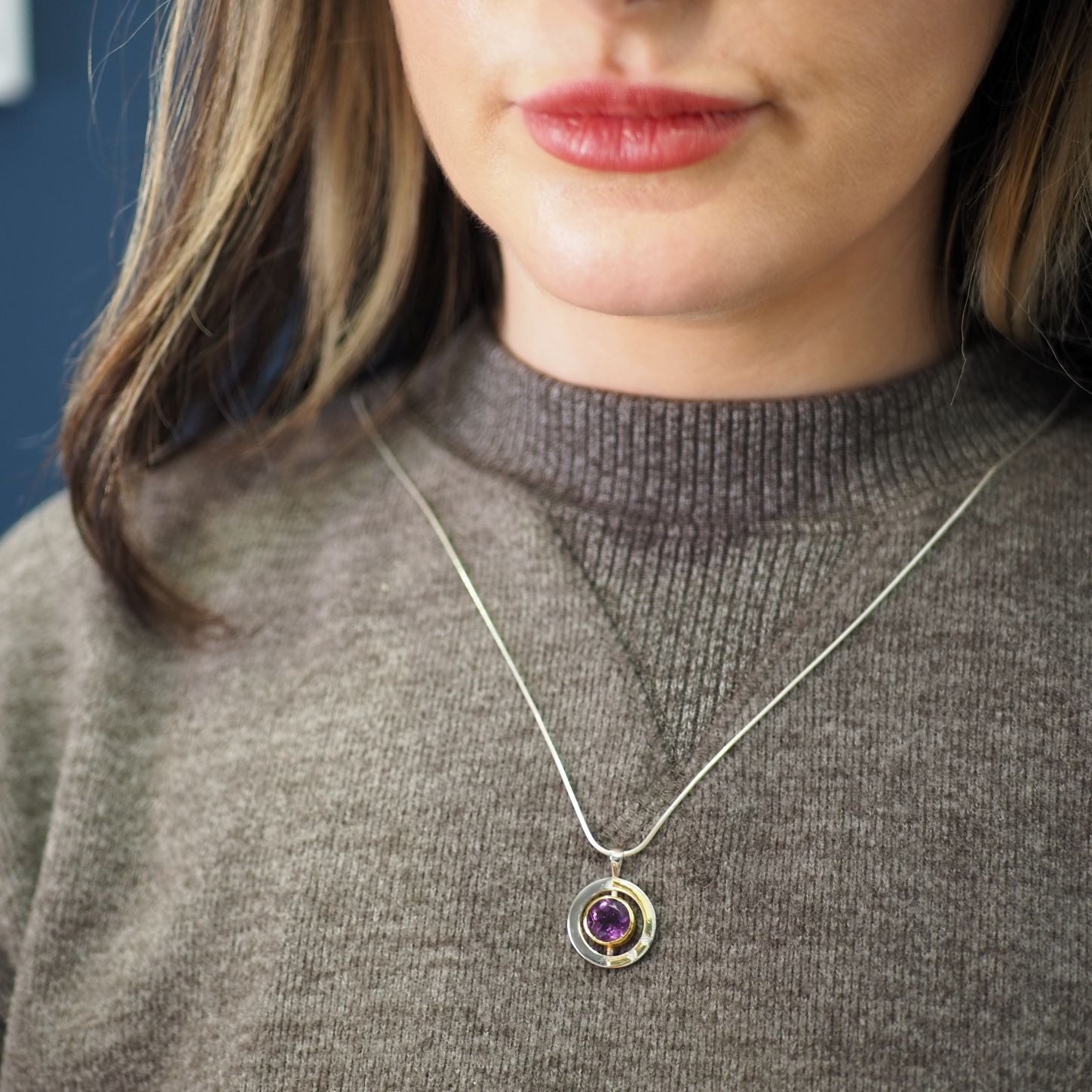 A person wearing a gray sweater and a silver necklace with a circular pendant that features a small purple gemstone. The focus is on the Elodie Pendant in silver gold & various gemstones by Gallardo & Blaine Designs, highlighting its intricate design and