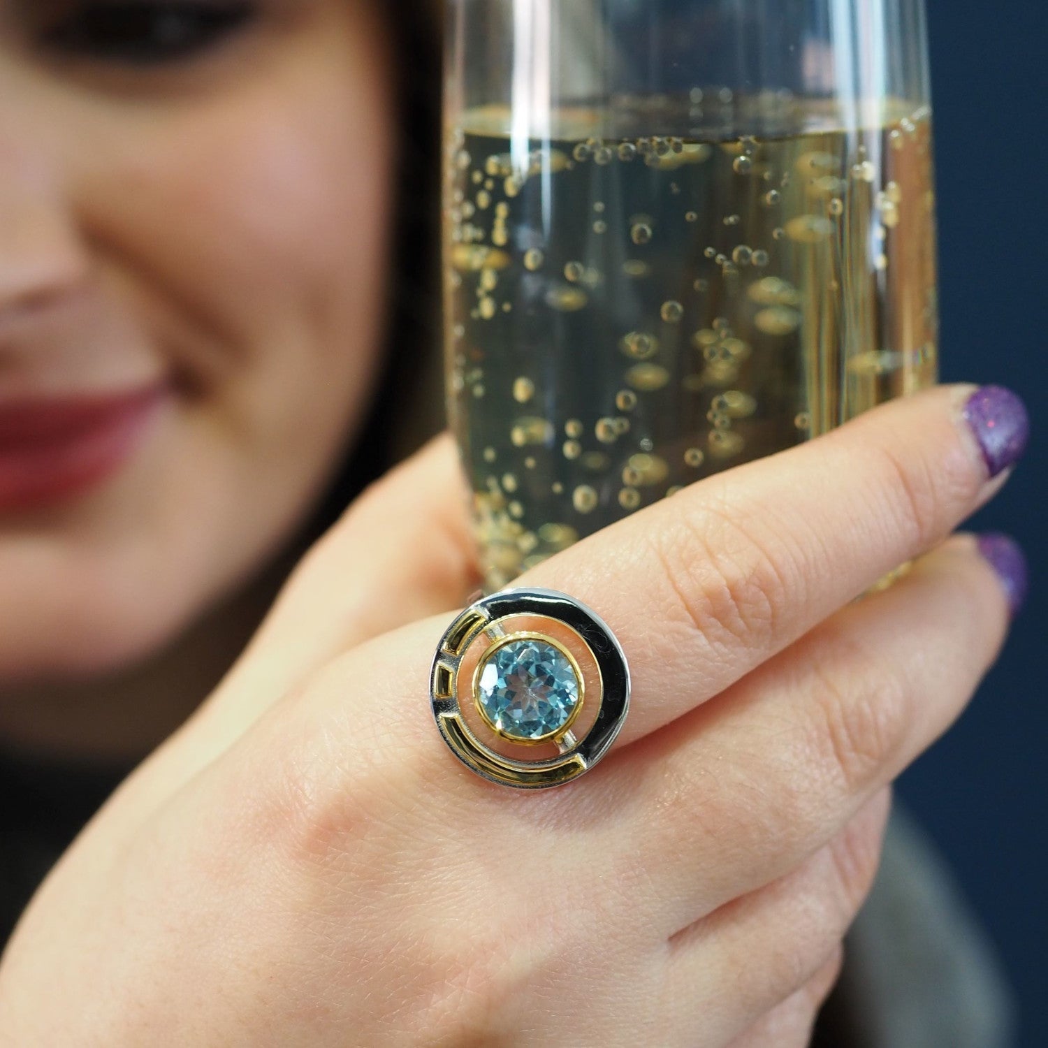Close-up of a person holding a glass of sparkling beverage. The focus is on their hand, adorned with the Gallardo & Blaine Designs Elodie Ring in Silver Gold & various gemstones, exuding vintage style glamour. The person's face is slightly blurred in the