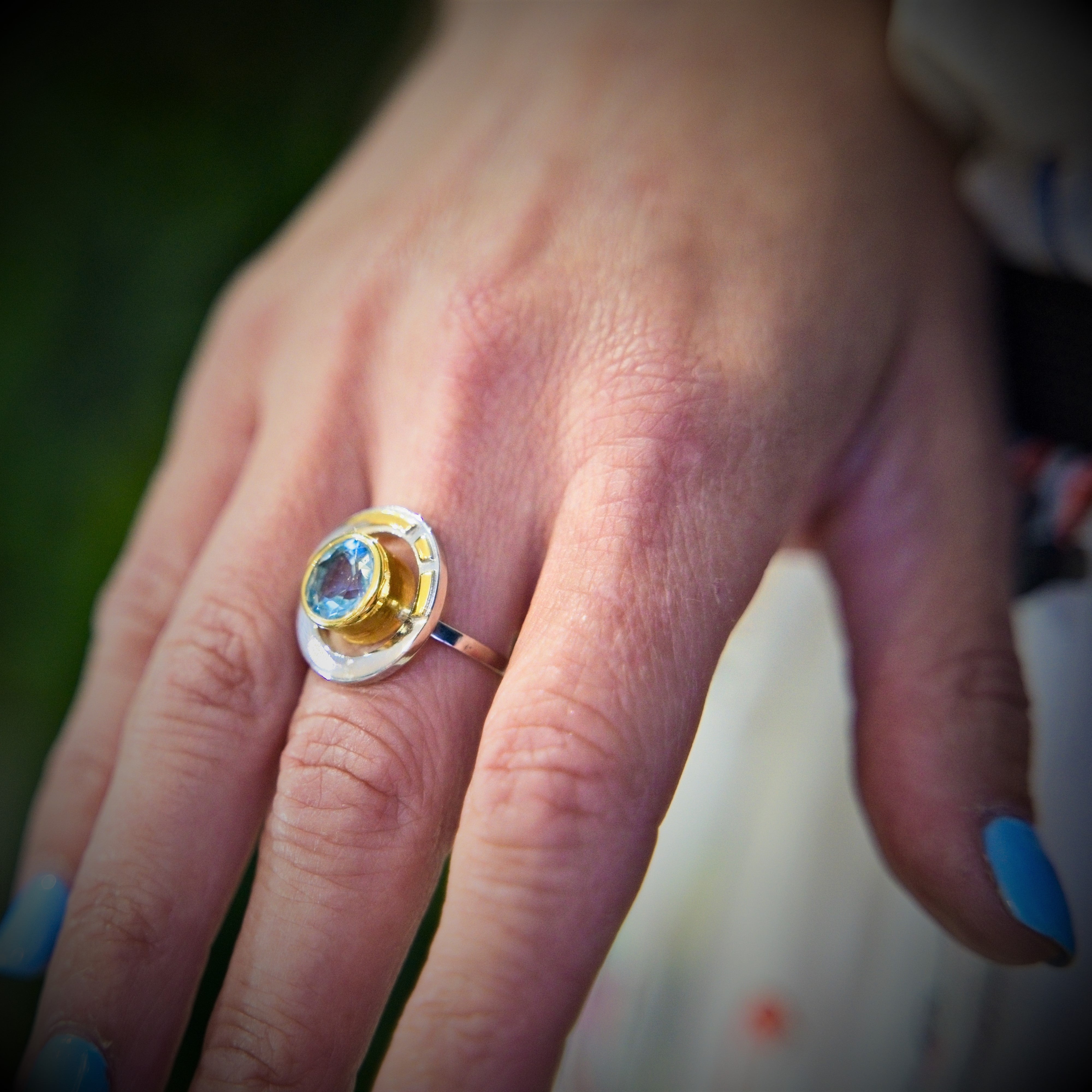 A close-up image of a hand with light blue nail polish wearing the Elodie Ring in Silver Gold & various gemstones by Gallardo & Blaine Designs. The ring features a yellow and silver band with a round blue gemstone at its center, exuding vintage style glam