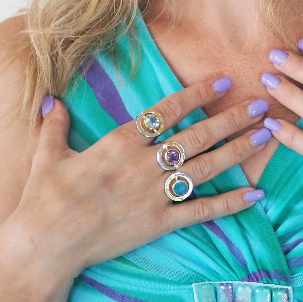 A person with long, light hair and lavender nail polish showcases vintage style glamour by wearing three gemstone rings on their fingers. The rings, including an elegant Elodie Ring in Silver Gold & various gemstones from Gallardo & Blaine Designs, featur