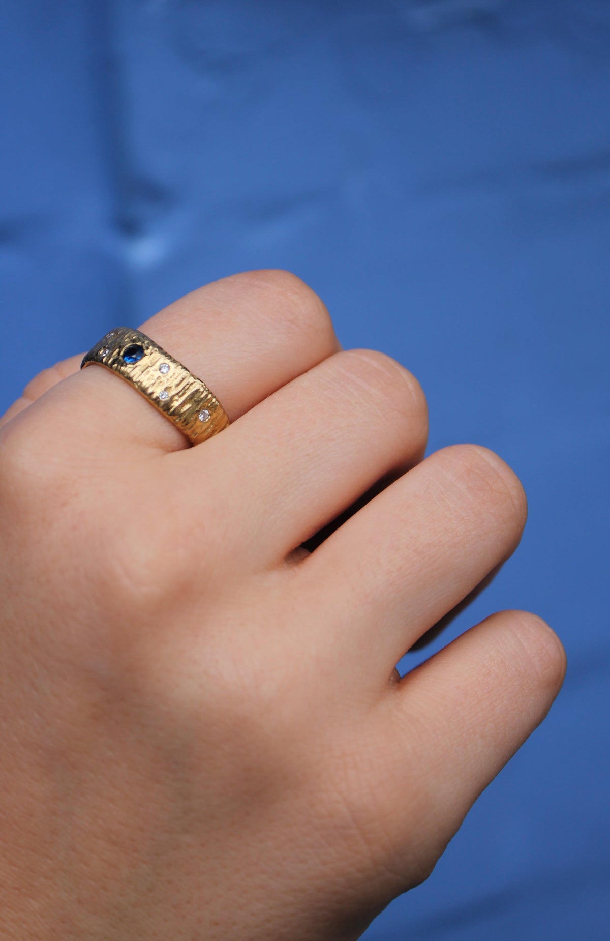 A close-up of a fair-skinned hand wearing the Etta Ring with Sapphire & Diamonds by Gallardo & Blaine Designs, featuring a 9ct yellow gold hammered band, set against a blue background.