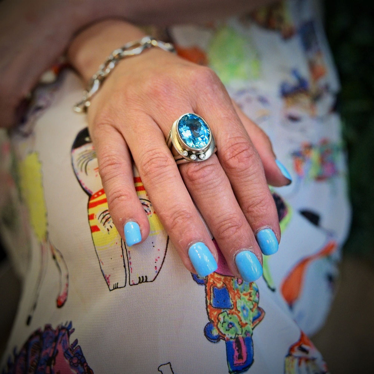 A close-up of a hand with blue nail polish, adorned with a statement Eyetelia Ring in various gemstones by Gallardo & Blaine Designs on the middle finger. The person is dressed in colourful, cat-patterned fabric, and an adjustable design silver bracelet g
