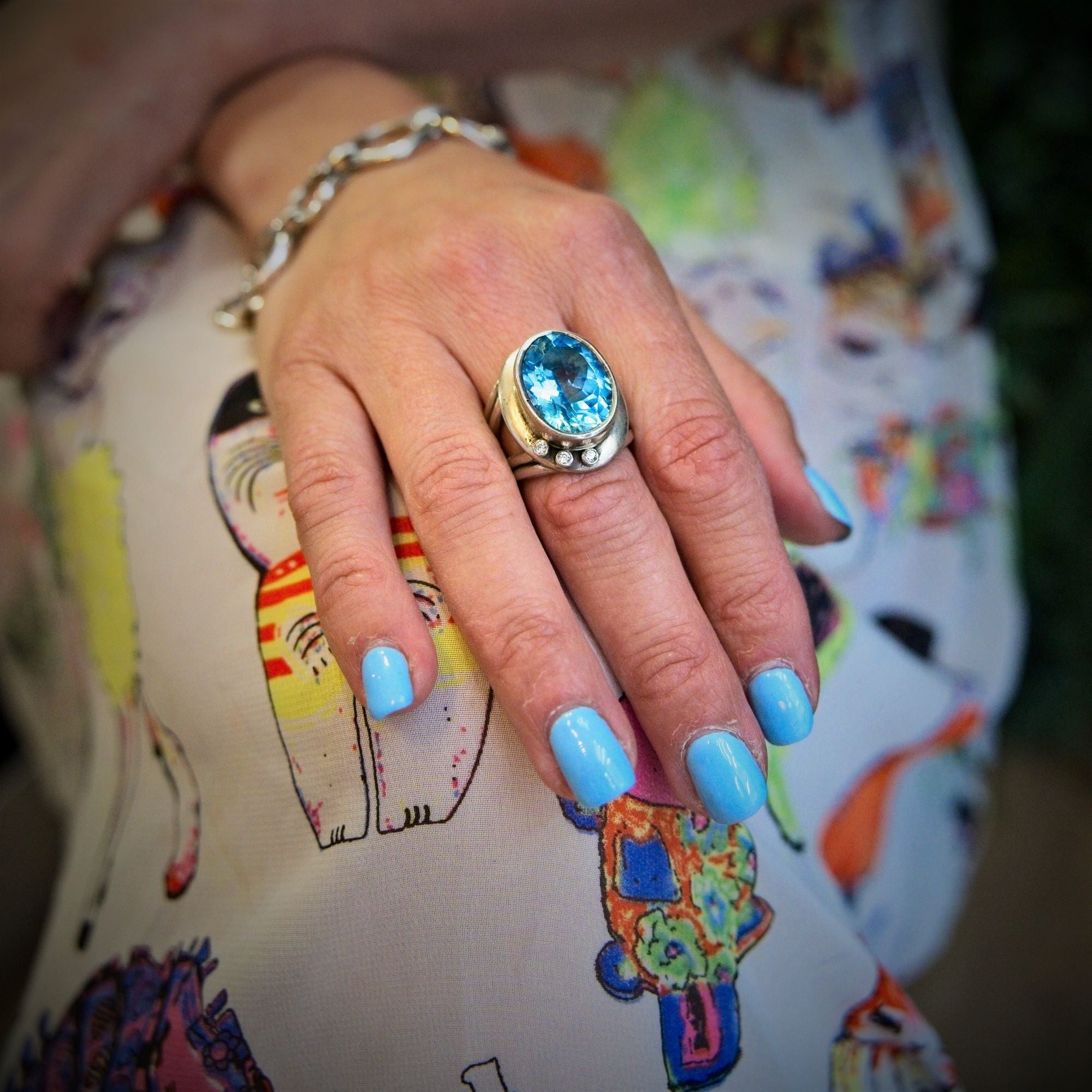 A hand with light blue nails wears the Gallardo & Blaine Designs Eyetelia Ring in blue gemstone, paired with a silver bracelet, resting on colorful cat-patterned fabric.