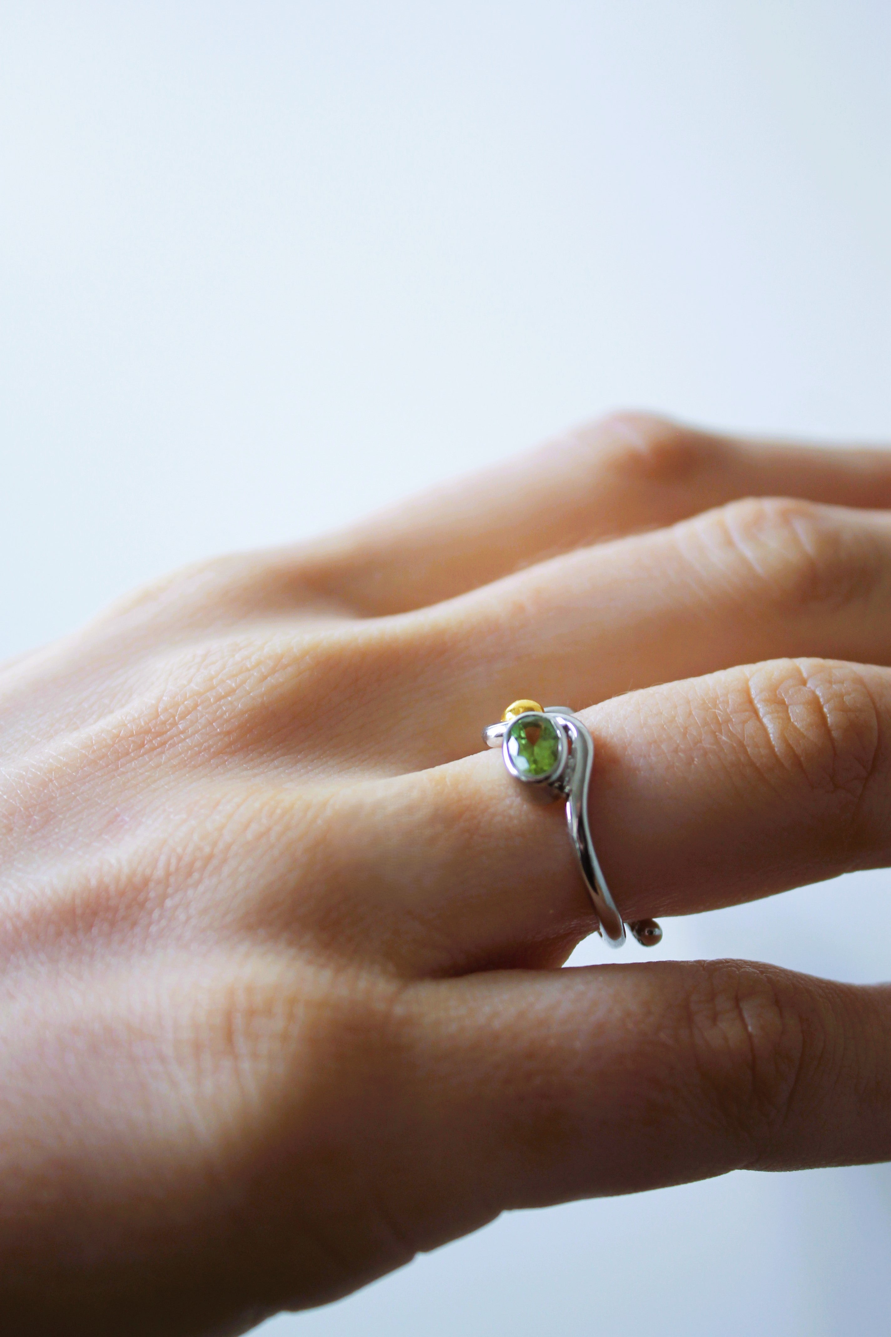 A close-up of a hand wearing Gallardo & Blaine Designs’ Adjustable Heartbeat Ring, crafted in sterling silver and featuring a small green gemstone, shown on the ring finger against a plain light background.
