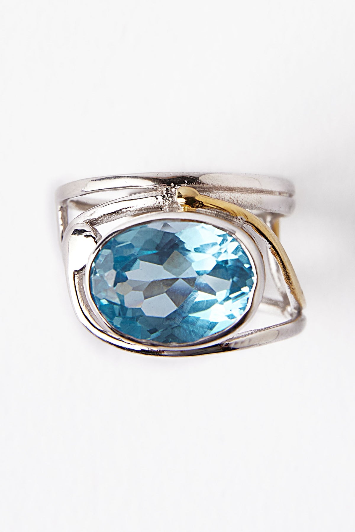 The Gallardo & Blaine Designs Horizontal Ring showcases a silver band with an oval faceted blue gemstone at its center, elegantly accented by a delicate gold curve on one side, creating a striking contrast against the plain white background.