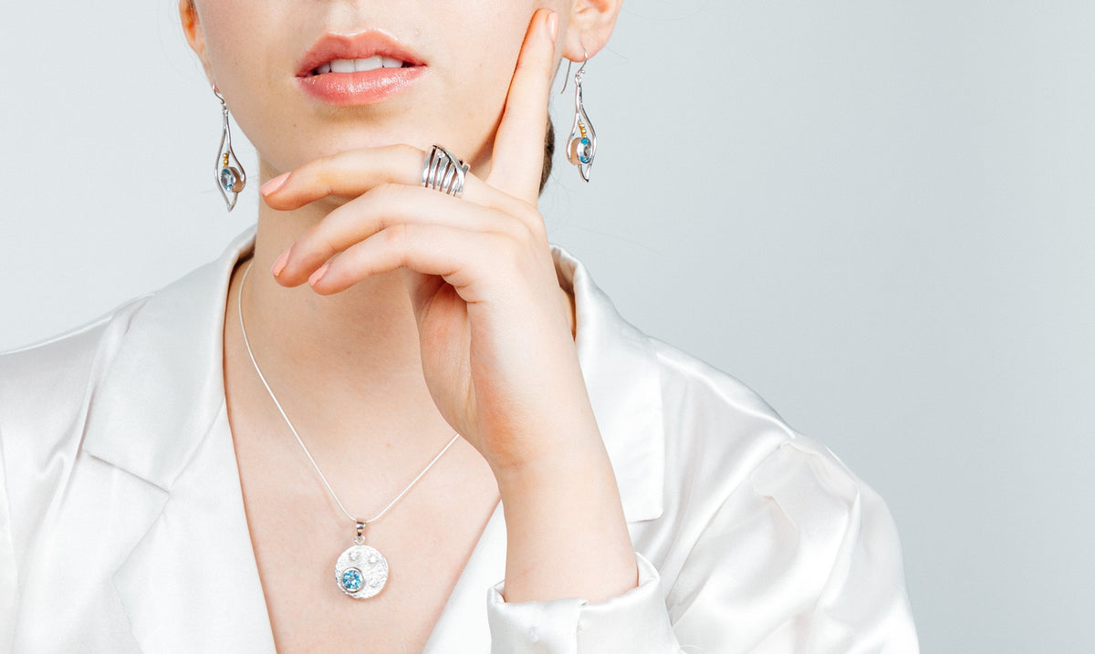 A person wearing elegant silver & gold jewelry, including Gallardo & Blaine Designs Iris Earrings in various gemstones, rings, and a necklace. The necklace features a round pendant with a blue gem. The person is dressed in a white satin blouse, with their