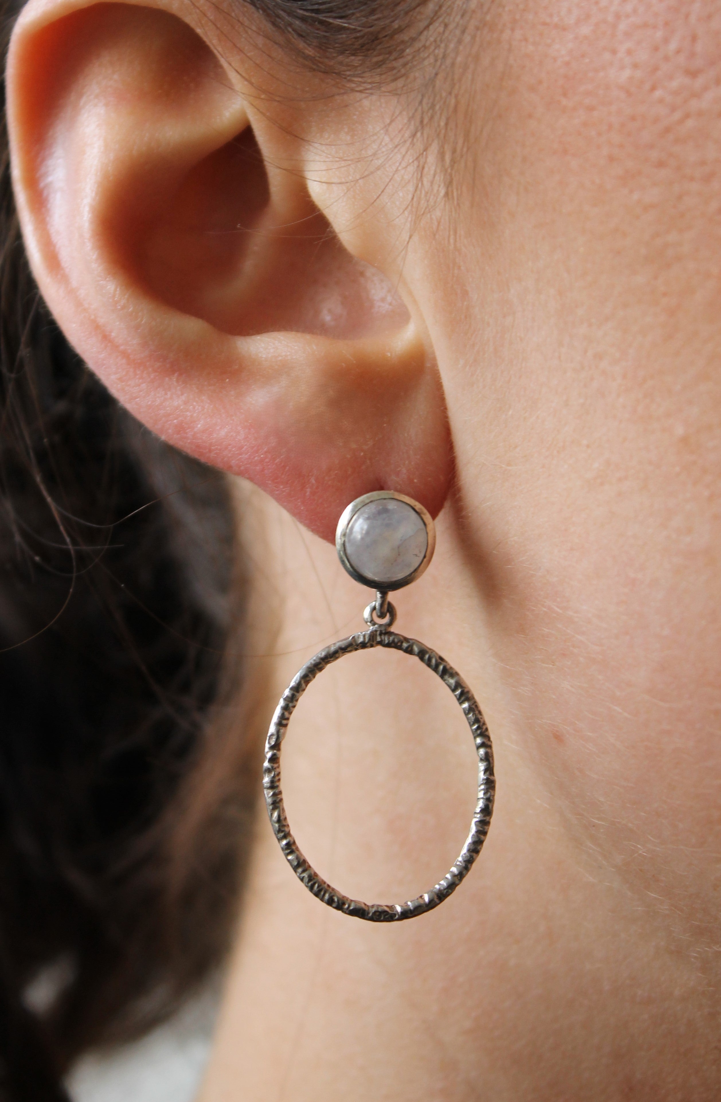 A close-up of a person’s ear wearing Gallardo & Blaine Designs’ Lunar Earrings, featuring various gemstones in a round stud with a large, textured circular hoop below.
