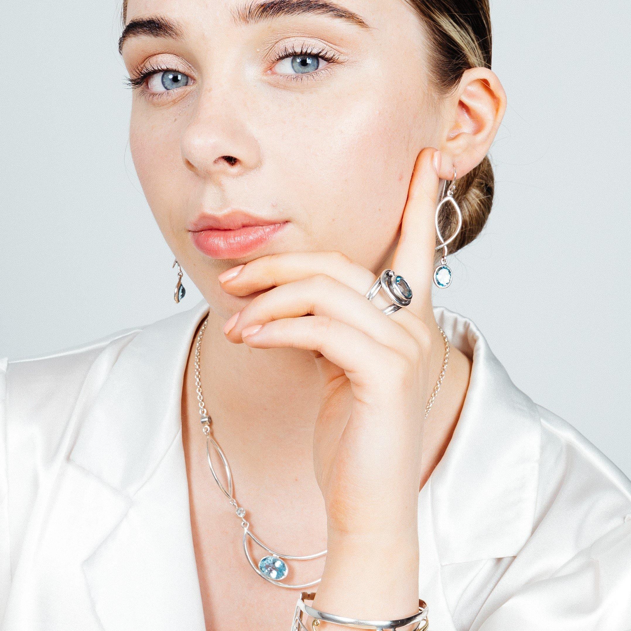 A woman with fair skin, blue eyes, and light brown hair is wearing a white satin blouse. She has on matching silver jewelry, including a necklace, rings, and a bracelet, all featuring blue gemstones. Her elegant drop silver earrings from Gallardo & Blaine