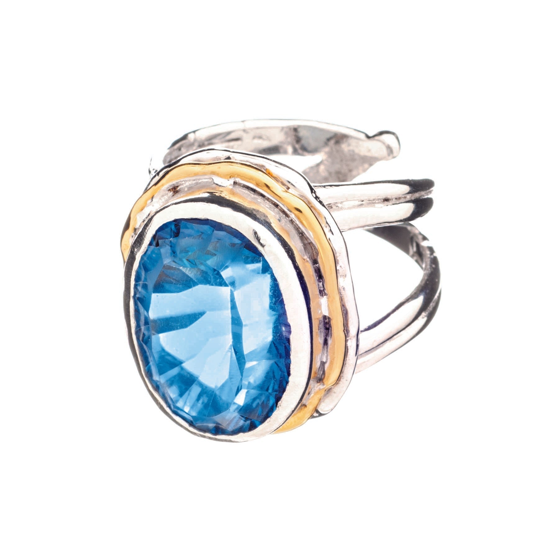 The Magnolia Ring in silver gold & various gemstones by Gallardo & Blaine Designs is a stunning statement piece crafted from sterling silver. It features a large, faceted blue gemstone as its oval-shaped centerpiece, accented by gold-toned bands that enha