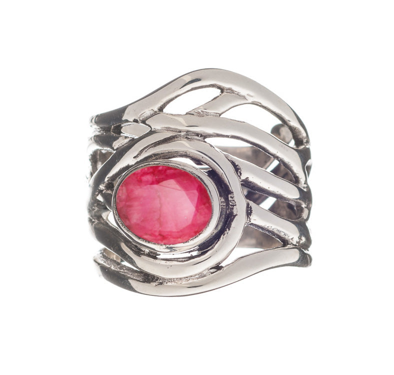 A silver, adjustable ring with an intricate, wavy band design features an oval-shaped, faceted pink gemstone set in the center. The metalwork creates an elegant and modern look, enhancing the vibrant color of the gemstone. This is the Mayhem Ring in vario