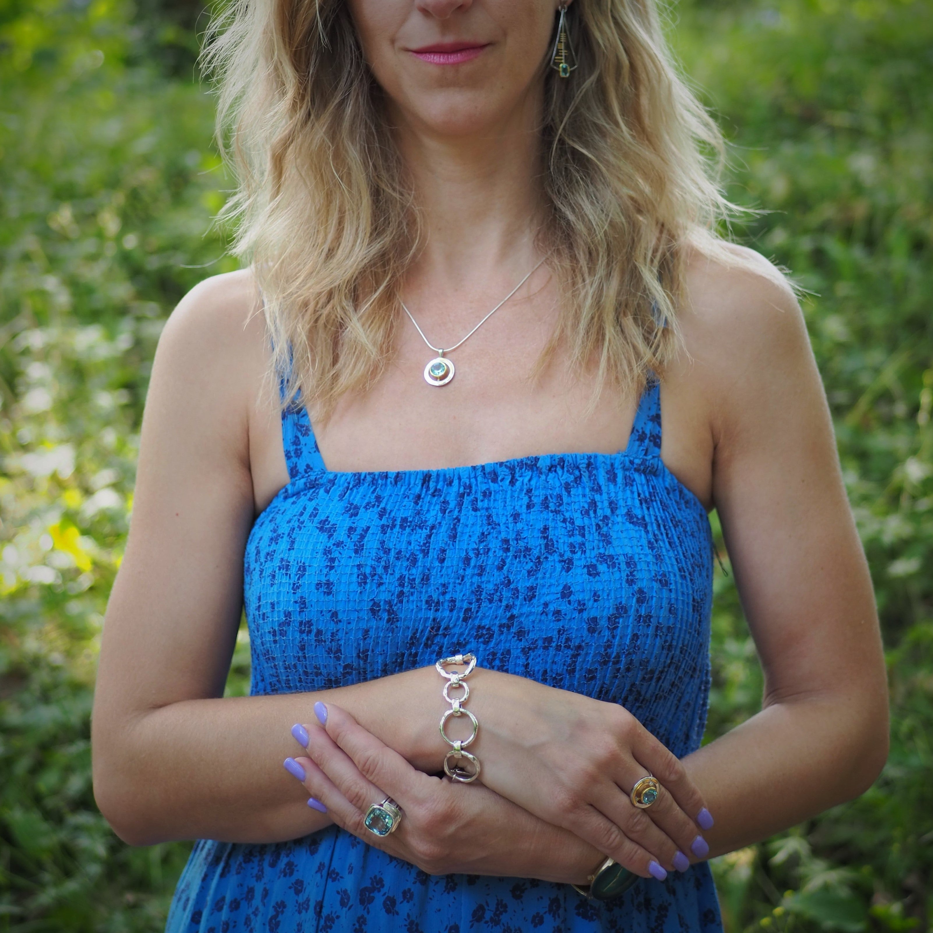A woman with wavy blonde hair wears a sleeveless blue dress and stands outdoors. She is adorned with jewelry, including earrings, a necklace, and a bracelet. Her hands are positioned in front of her, showcasing an exquisite Art Deco Ring in Silver Gold &