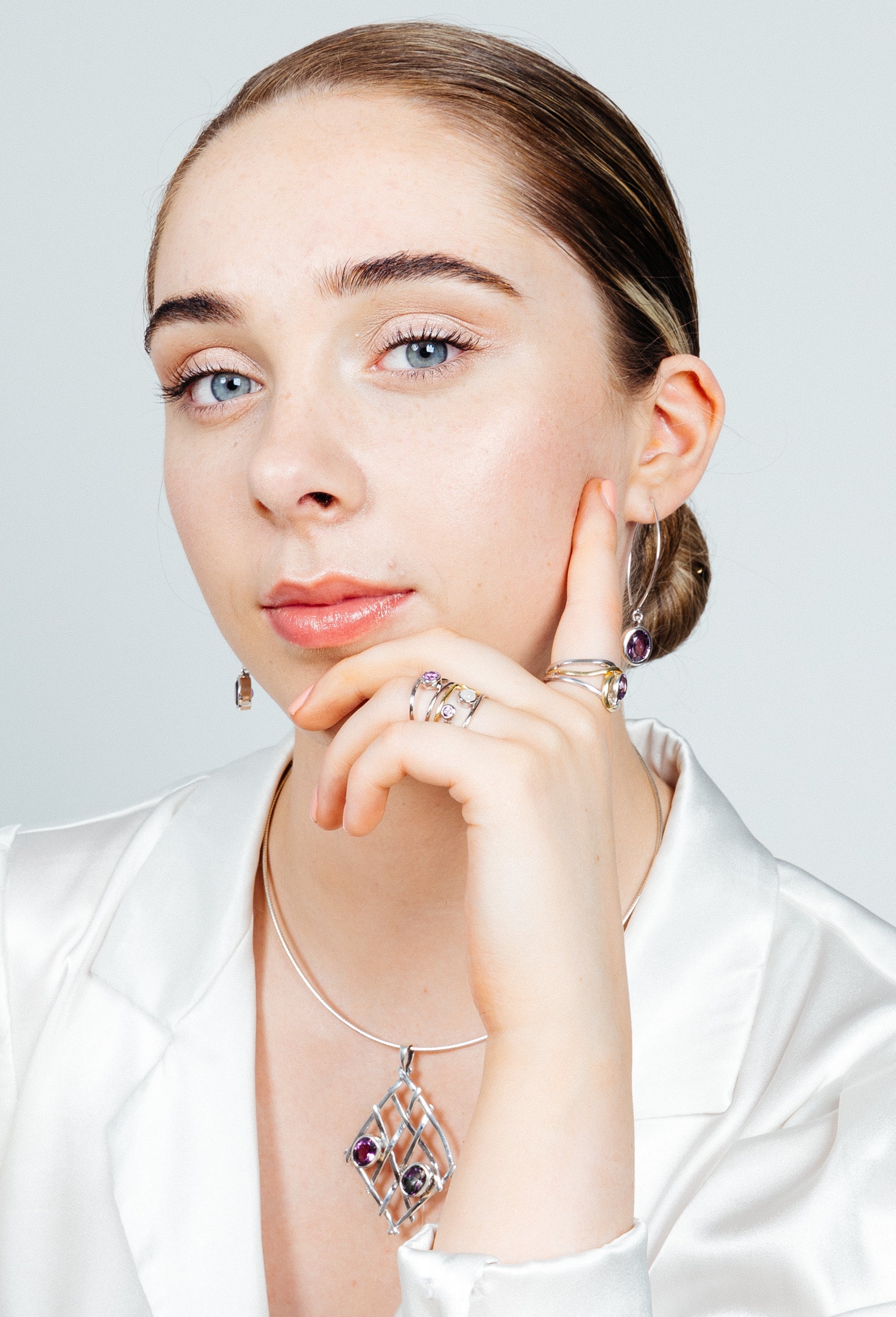 A blue-eyed individual in a white satin top poses with a hand on their chin, showcasing Gallardo & Blaine Designs' Nettle Pendant in various gemstones. They wear matching sterling silver earrings and rings with purple stones against a plain white backgrou