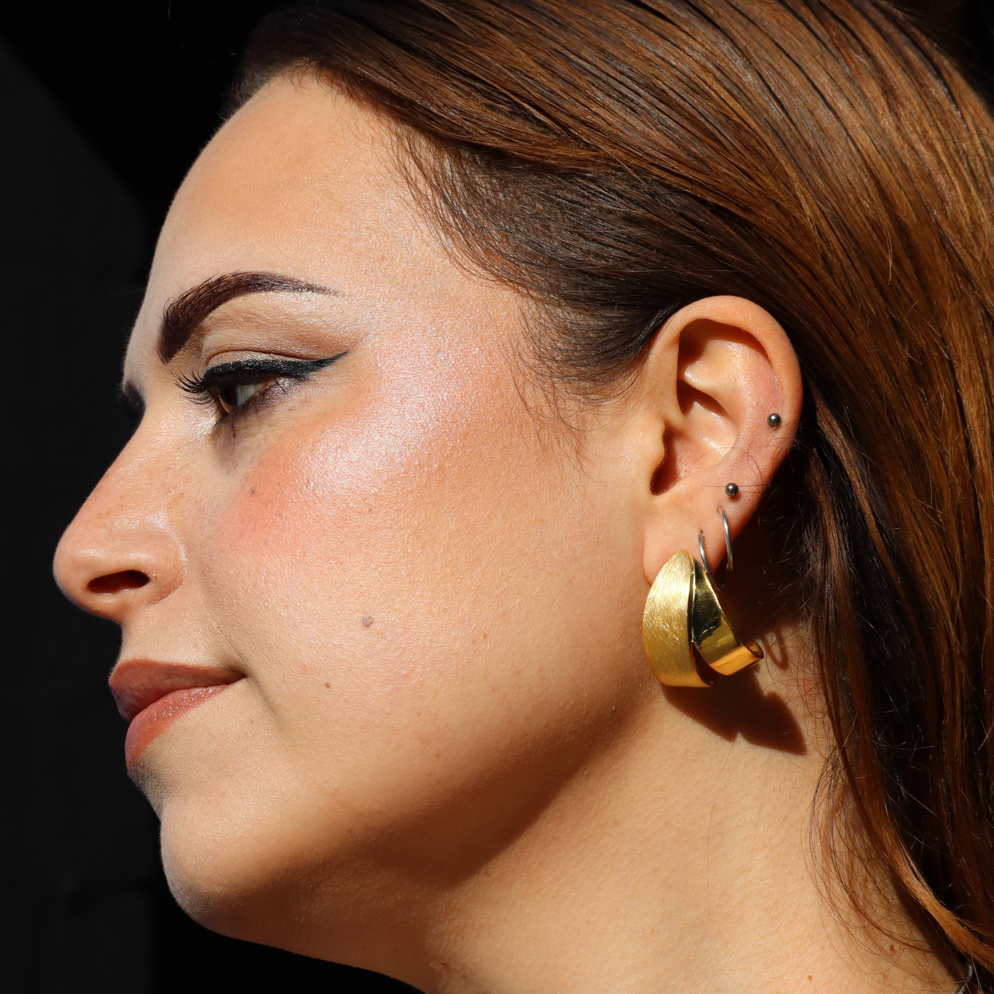 A woman with light brown hair, wearing Gallardo & Blaine Designs Origata Studs and multiple small earrings, is shown in profile with winged eyeliner and natural makeup, highlighted by sunlight.