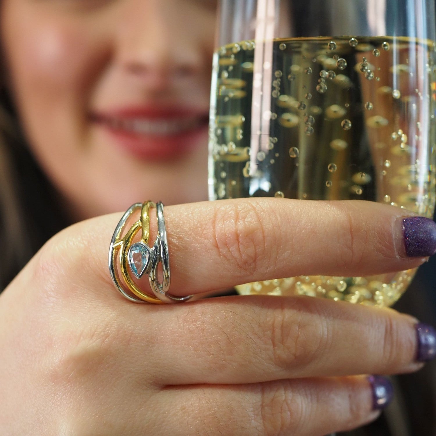 A person with long hair is holding a glass of sparkling wine. They are wearing an elegant, dainty Peacock Ring in various gemstones by Gallardo & Blaine Designs, adjustable in silver and gold, and have purple nail polish. The background is slightly blurre