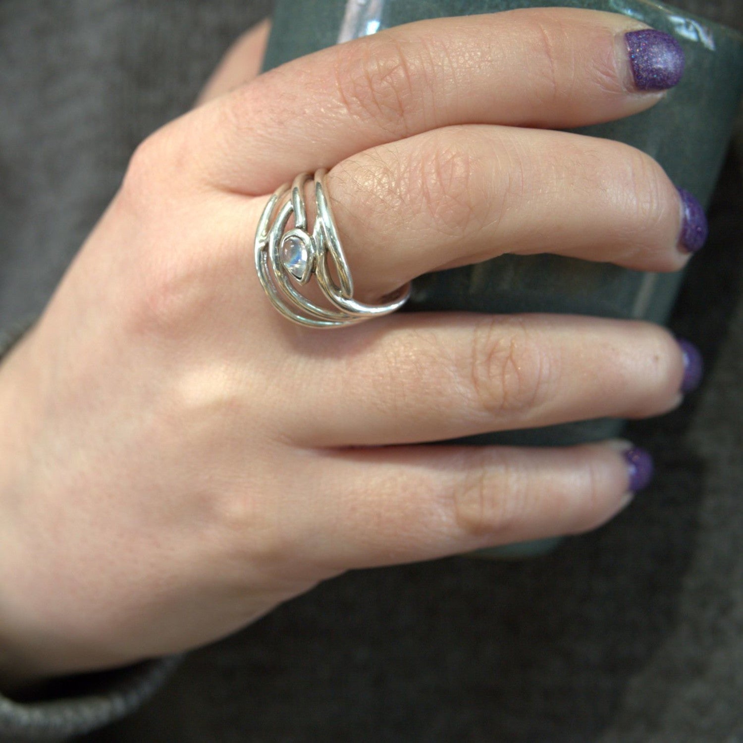 Close-up of a hand holding a greenish-blue ceramic cup. The hand is adorned with an elegant dainty ring featuring an intricate, wavy design and a central gemstone. This beautiful ring, called the Peacock Ring in various gemstones by Gallardo & Blaine Desi