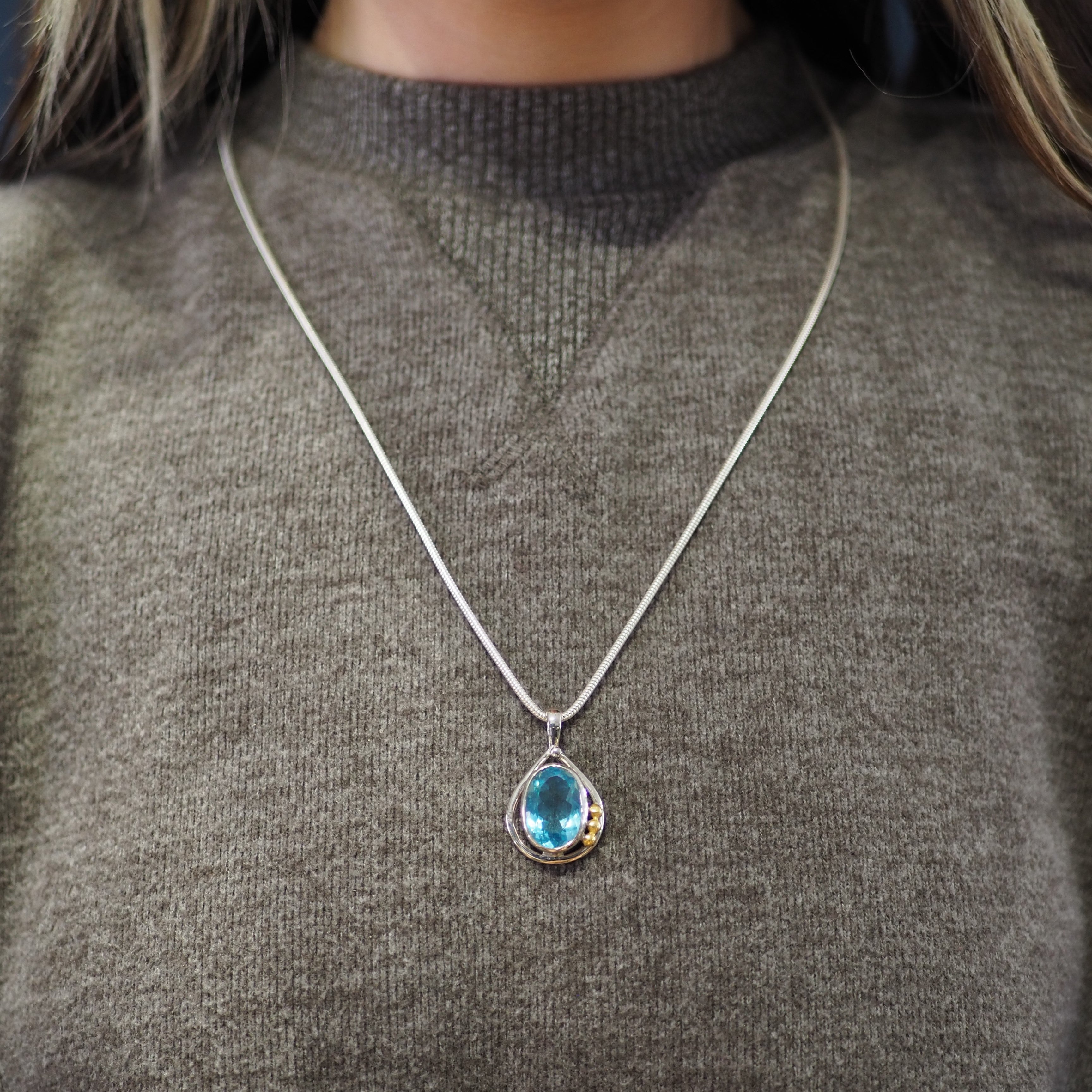A person wearing a gray sweater has a sterling silver necklace with a teardrop-shaped blue gemstone pendant from Gallardo & Blaine Designs called the Poppy Pendant Small in various gemstones. The background is blurred, focusing on the necklace and sweater