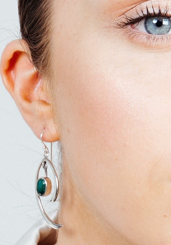 Close-up shot of a person wearing minimalist elegance with a pair of Primrose Earrings in various gemstones by Gallardo & Blaine Designs. The sterling silver earrings feature an oval green gemstone and modern, artistic design with curved silver elements.