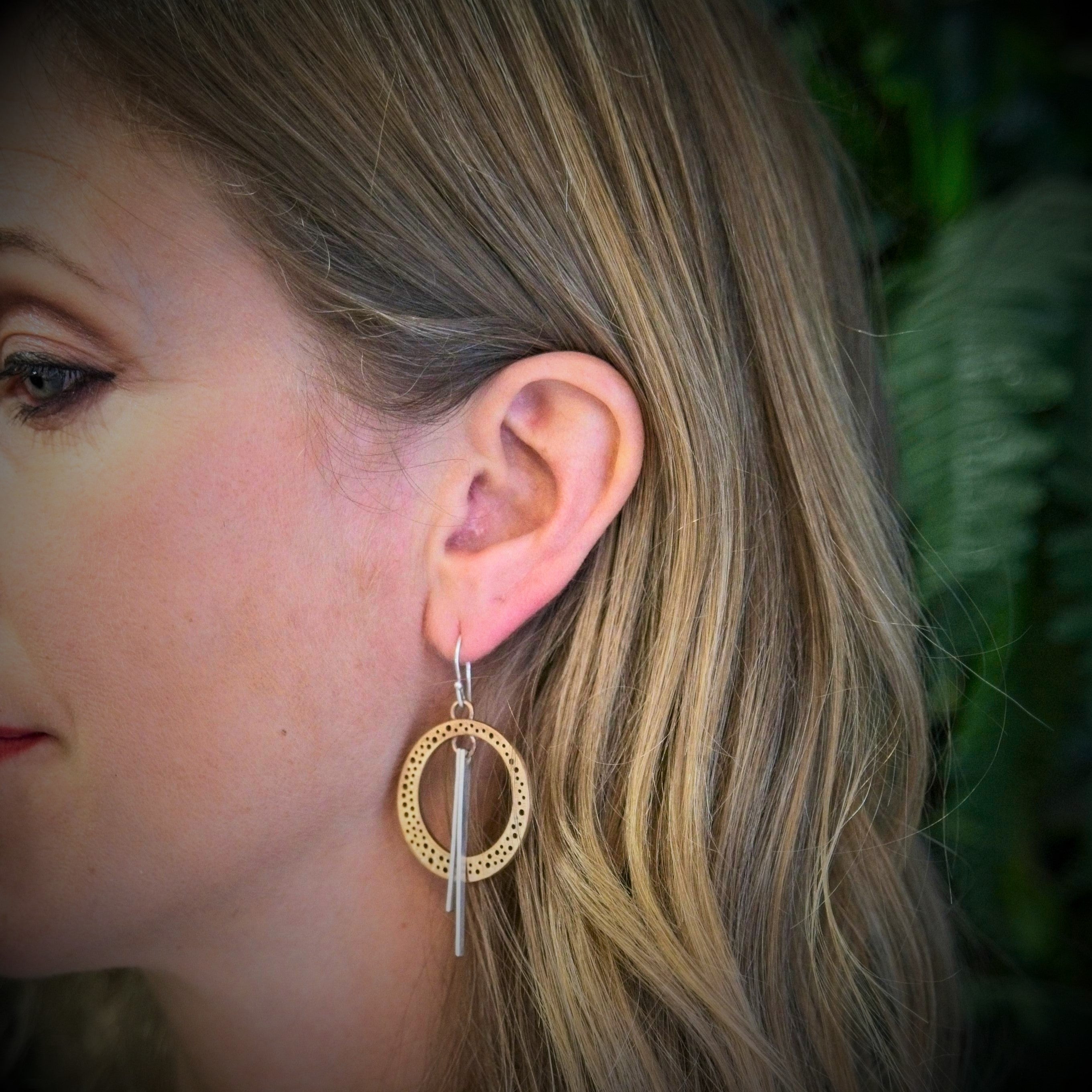 A close-up of a woman with light brown, wavy hair wearing Gallardo & Blaine Designs' Rivera Earrings in gold with a vertical sterling silver bar. The large, circular Art Deco jewelry is set against blurred green foliage.