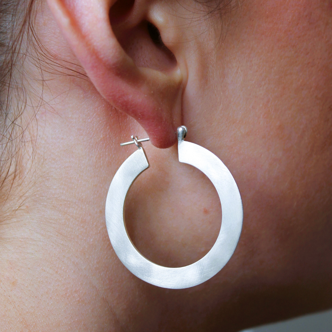 Close-up of a person wearing Gallardo & Blaine Designs’ Satin Hoop Earring in silver—large, round with a brushed metal finish and flat surface, worn through a pierced earlobe.