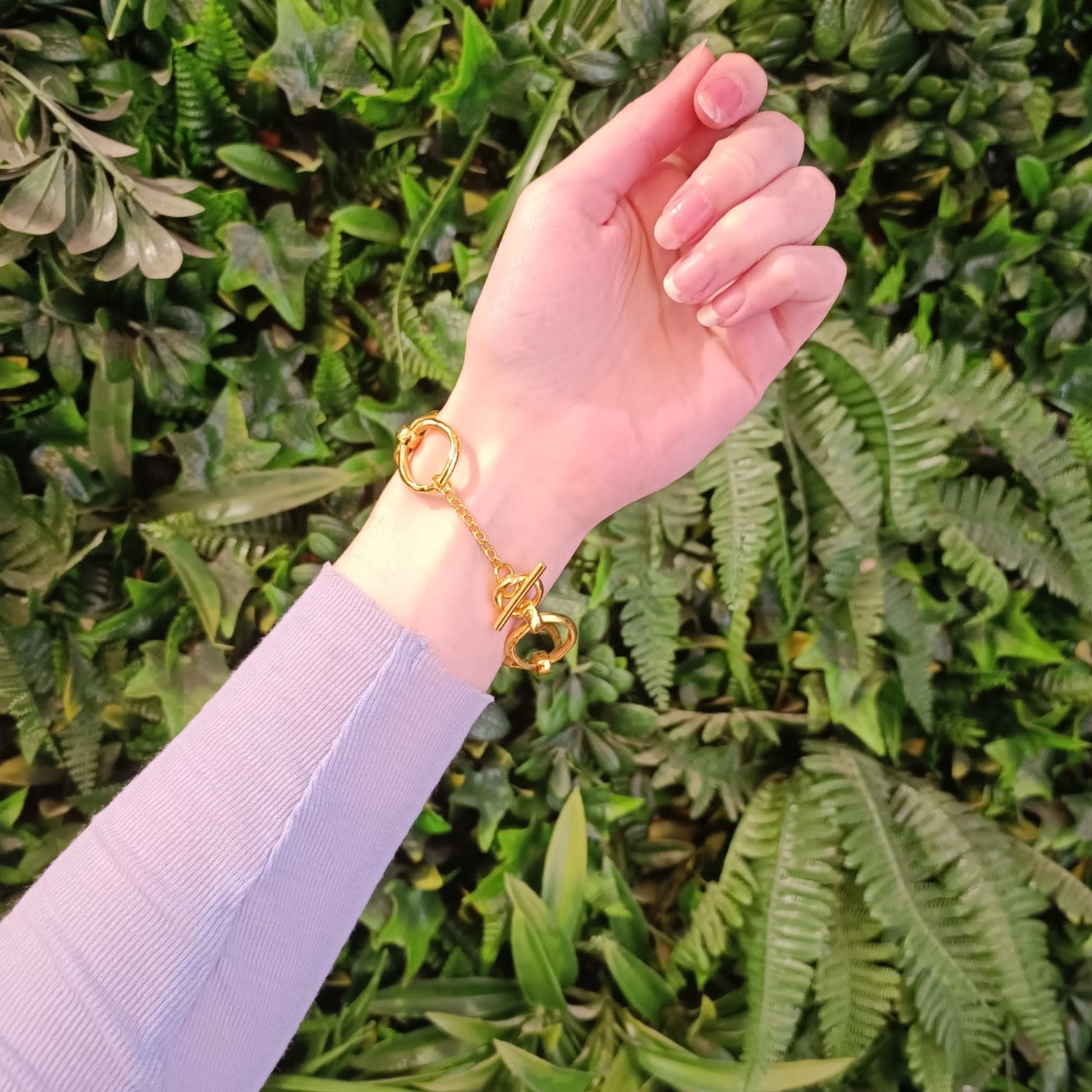 A Sea Honey Bracelet Gold Plated Bronze on a person's wrist against a background of greenery. The gold-colored bracelet glimmers like charm jewelry, and the person is wearing a light purple long-sleeve shirt. The lush greenery consists of various types of