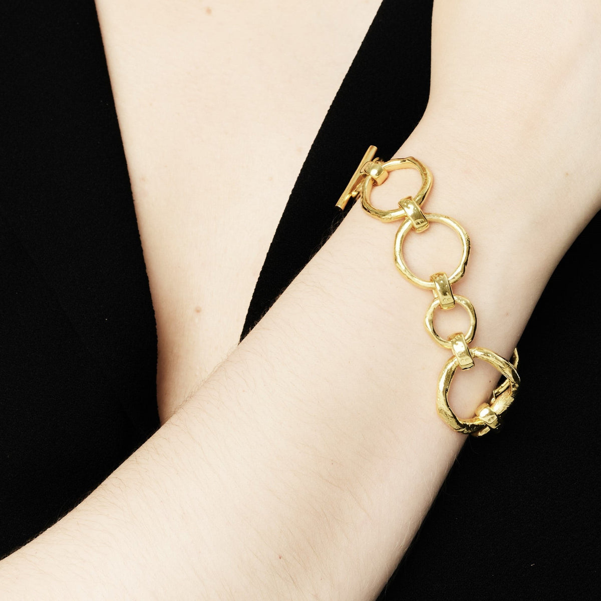 Close-up of a person wearing a black outfit showcasing a bracelet on their wrist. The accessory features large, interconnected circular bronze links with gold plating, adding a stylish touch to the elegant attire. The background is plain, emphasizing the