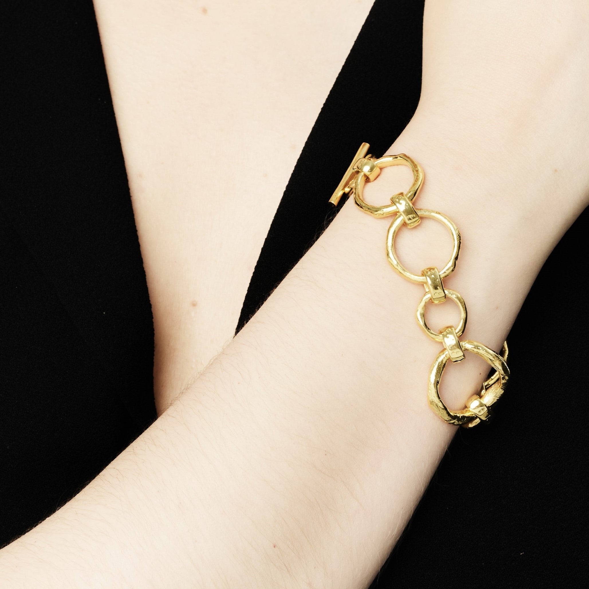 Close-up of a person wearing a black outfit showcasing a bracelet on their wrist. The accessory features large, interconnected circular bronze links with gold plating, adding a stylish touch to the elegant attire. The background is plain, emphasizing the