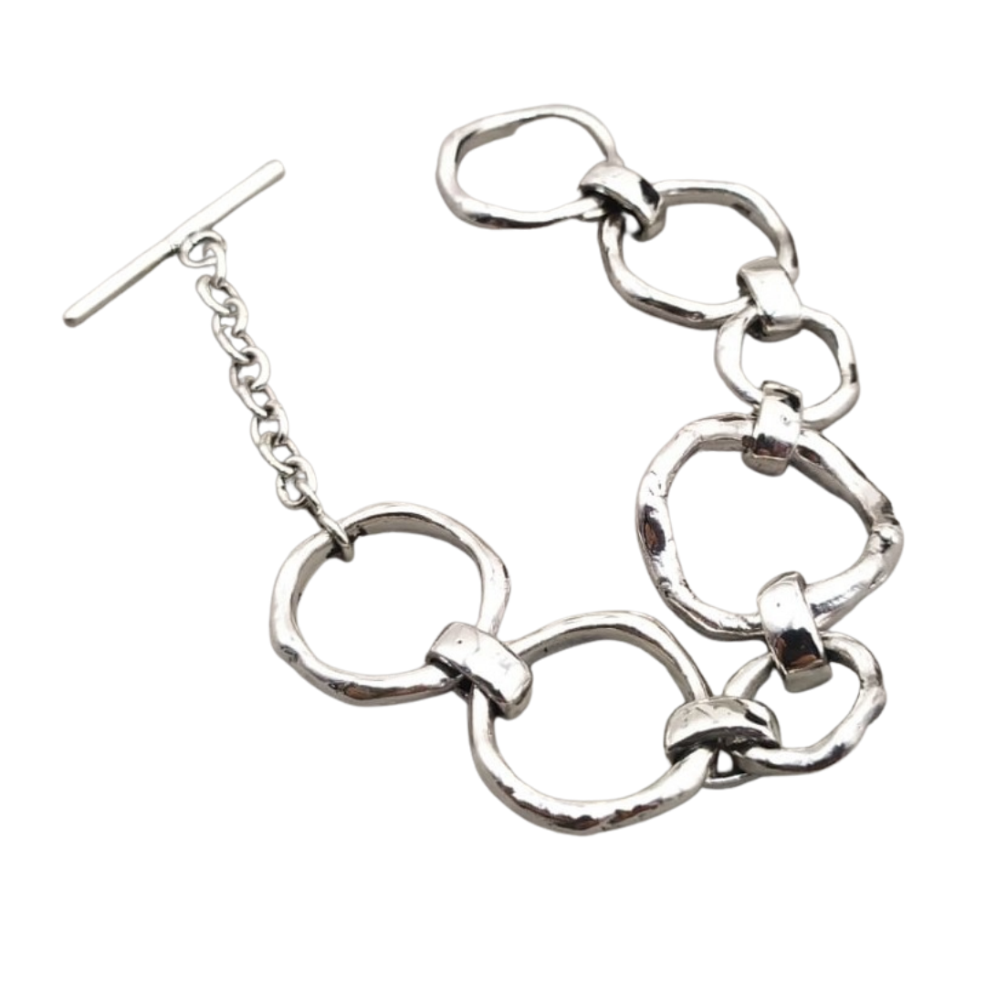 A stylish Sea Honey Bracelet Silver made of linked, thick silver rings is laid flat on a white background. It features a T-bar clasp with an adjustable chain, giving it a modern and elegant appearance. The metal has a slightly hammered texture, adding a t