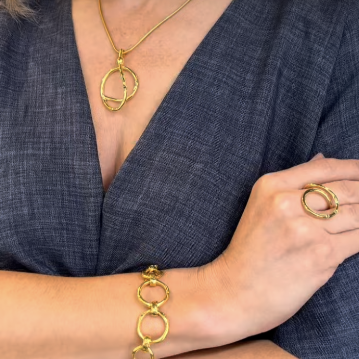 A person in a dark blue textured dress showcases gold jewelry: Gallardo & Blaine Designs' Sea Honey Necklace, a matching bracelet, and a gold ring with a circular design on their right hand.