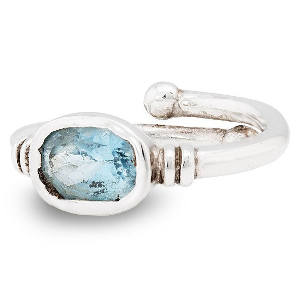 A close-up image of a silver adjustable ring featuring a rustic, oval-shaped blue gemstone. The setting has a raw and handmade appearance, with the stone secured by a simple bezel and adorned with subtle metal accents on the band. This is the Senna Ring i