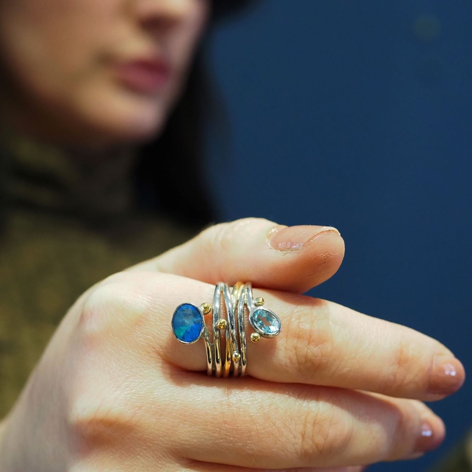 A close-up of a hand wearing several adjustable silver & gold rings with blue gemstones, including a striking Snake Ring in Silver Gold & various gemstones by Gallardo & Blaine Designs. The person’s face is blurred and partially visible in the background