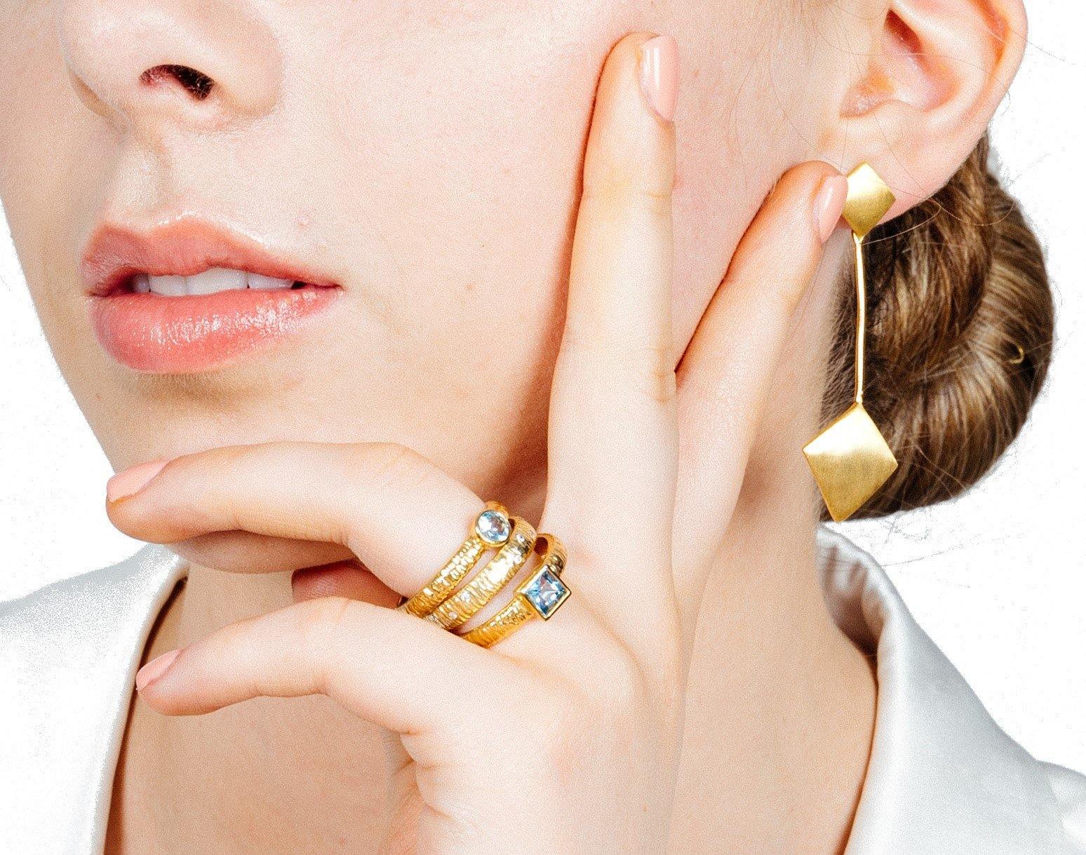 Close-up of a woman’s face and hand. She is wearing gold jewelry: two gold rings with gemstones on her fingers and an Art Deco Earrings In Gold Vermeil by Gallardo & Blaine Designs featuring a geometric design in gold vermeil. She is touching her face wit