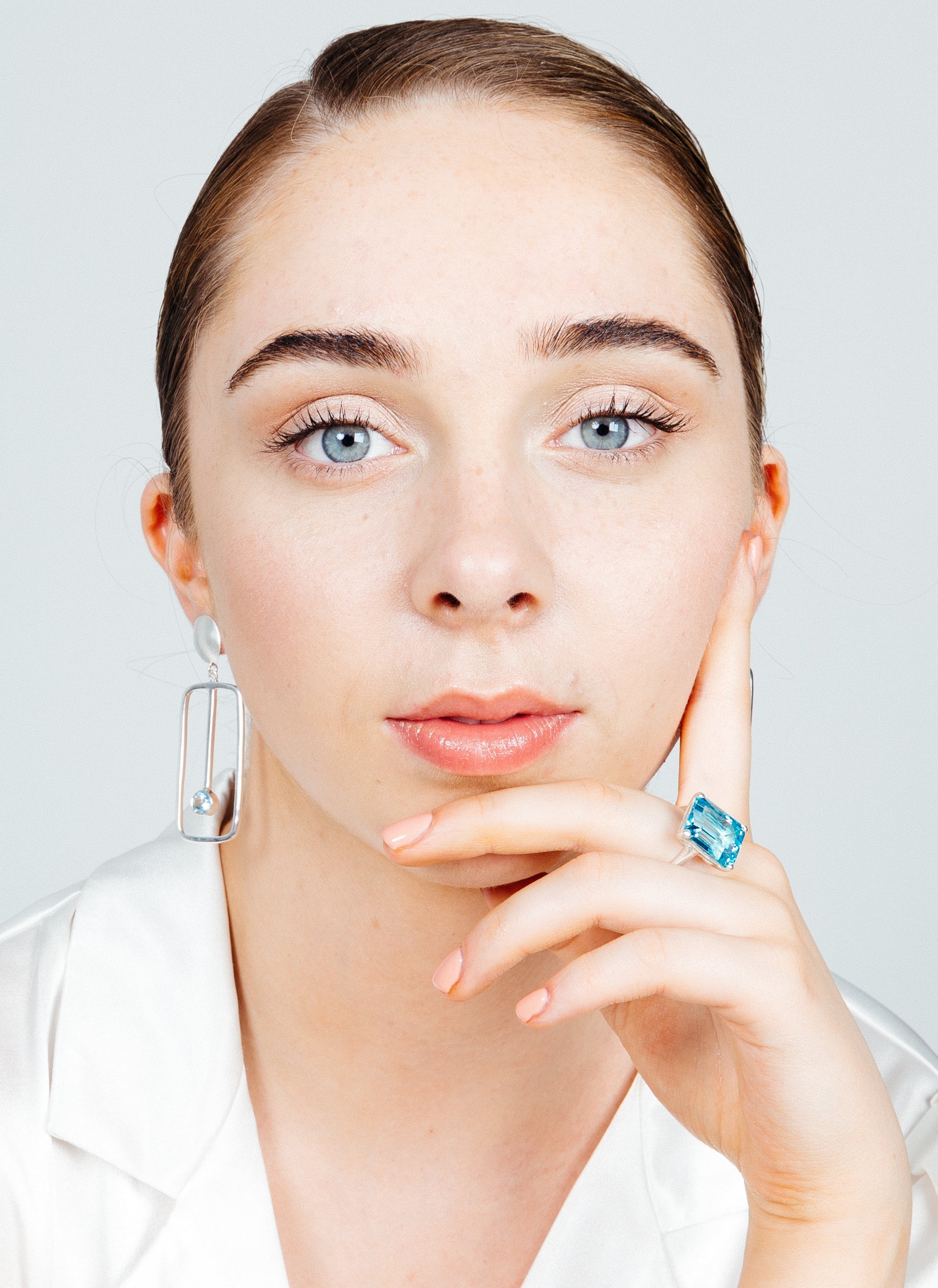 Close-up of a woman with light skin and blue eyes, staring directly at the camera. She has her hair neatly pulled back and rests her chin on her hand, adorned with a Gallardo & Blaine Designs Stellar Ring in Hydro Blue Topaz. She is wearing a white top an