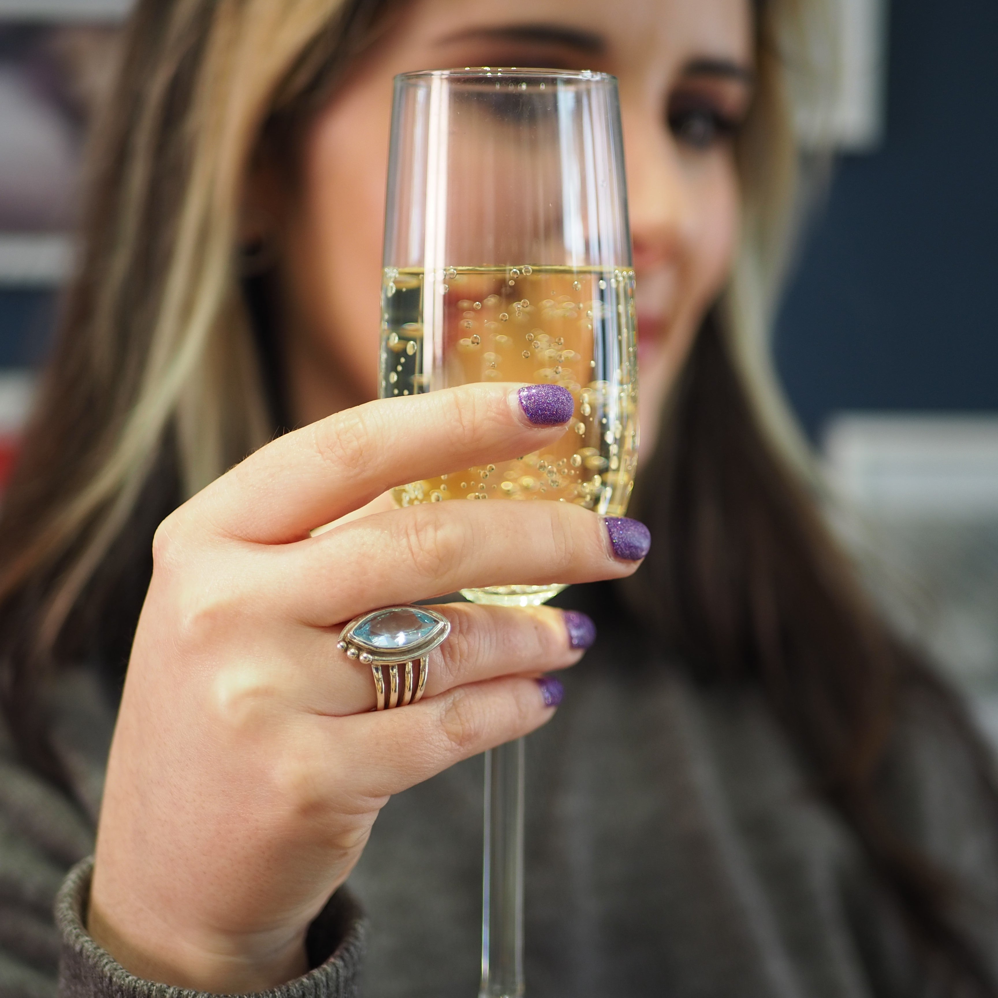 A person with long hair is holding a champagne glass filled with a bubbly drink. They have purple nail polish and are wearing the Tulip Ring in various gemstones by Gallardo & Blaine Designs on their finger. The background is blurred, focusing attention o