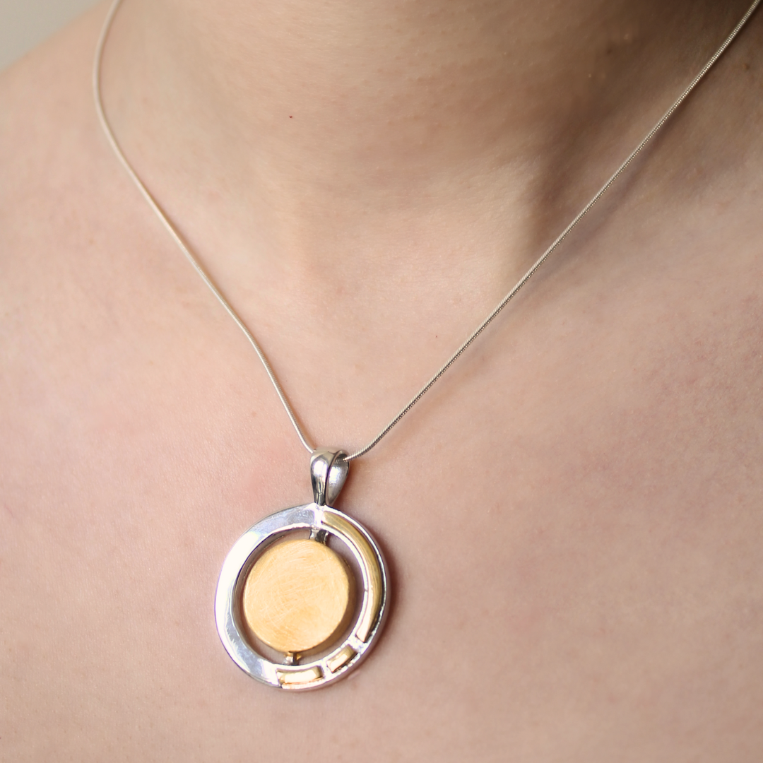 A close-up of the Urú Large Pendant & Chain by Gallardo & Blaine Designs, a silver necklace with a gold vermeil Celtic circular pendant featuring a gold-colored center, worn around a person’s neck.
