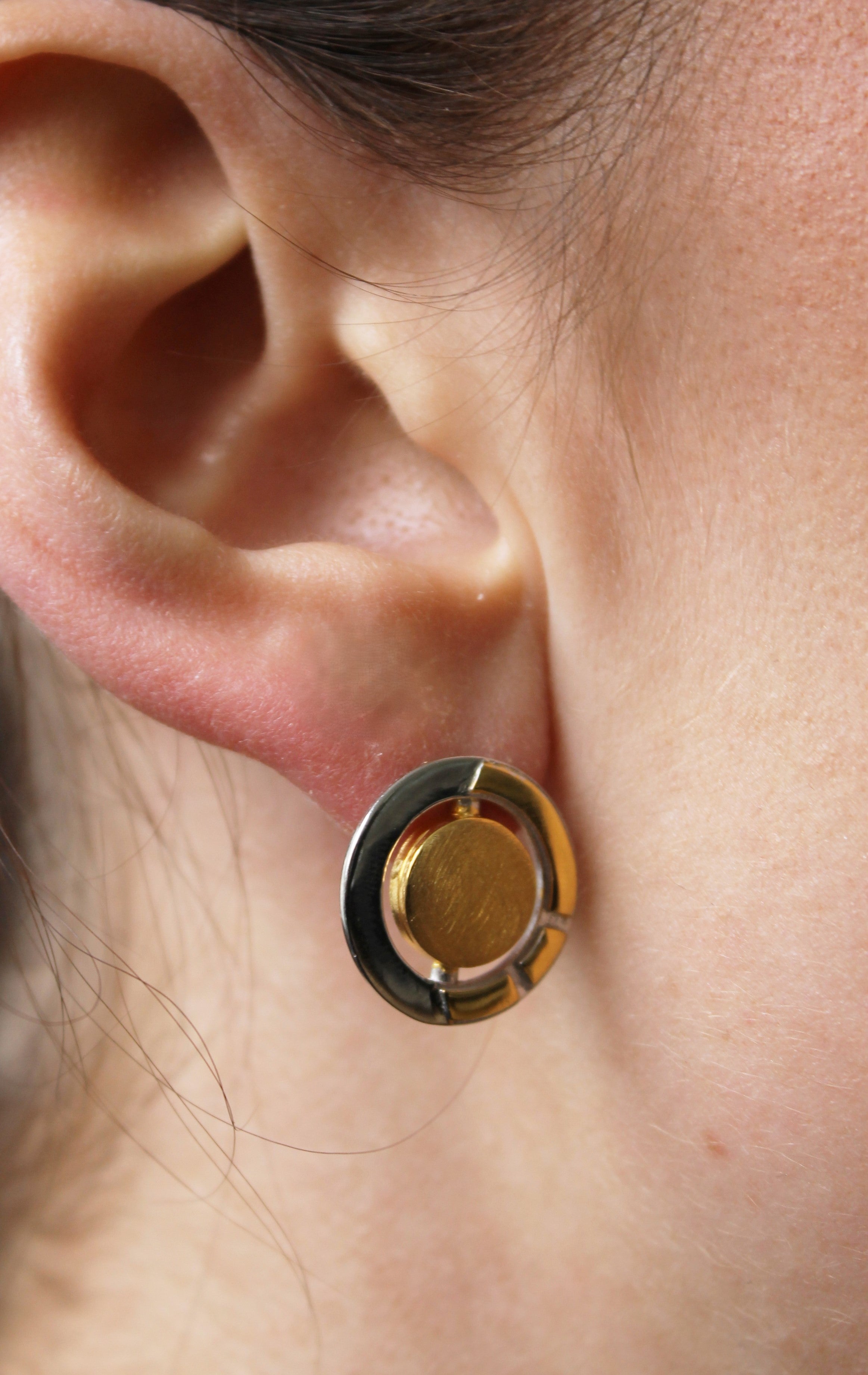 Close-up of a person's ear wearing the Urú Studs by Gallardo & Blaine Designs—round, gold and sterling silver geometric earrings inspired by Celtic motifs with a central gold circle encircled by a sterling silver design.