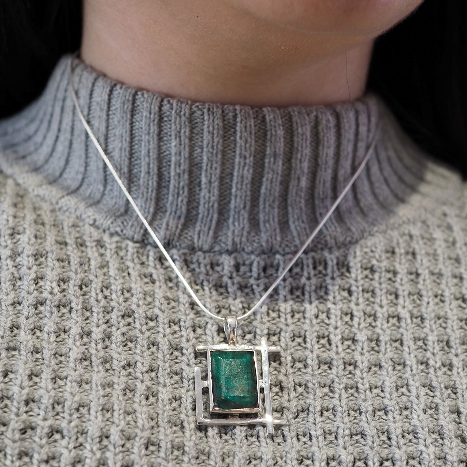 A person wearing a gray, ribbed, high-neck sweater with a sterling silver necklace featuring an Art Deco Pendant in various gemstones by Gallardo & Blaine Designs. The rectangular green stone is set in a geometric silver frame, hanging just below the coll