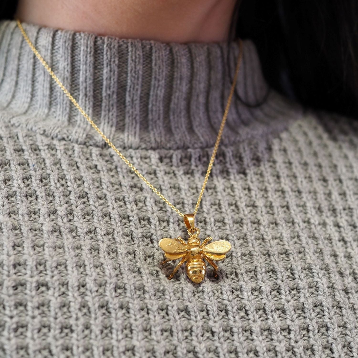 A person wearing a gray ribbed sweater and the Watermelon Tropical Wildlife Pendant - Large Bee With Chain—a gold-plated necklace perfect for fans of nature-inspired jewelry.