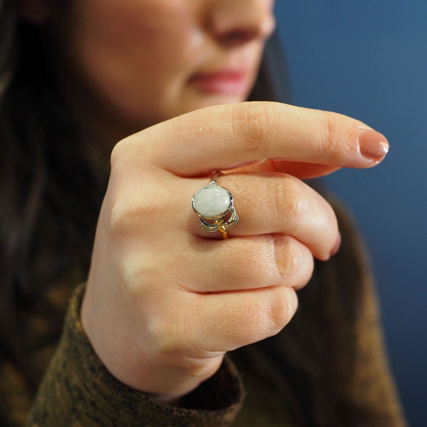 A person holds their hand up, displaying a delicate and dainty Daydream Ring in various gemstones with a round, opaque white stone set in a silver band by Gallardo & Blaine Designs. The person's face is partially visible, focusing on the timeless piece. T