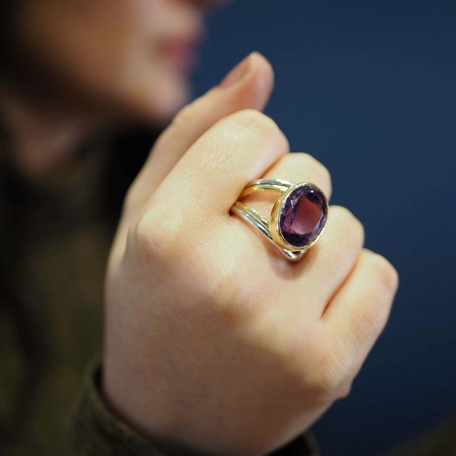 A close-up of a person's hand wearing the Dewberry Ring in various gemstones by Gallardo & Blaine Designs, featuring a chunky statement ring with a large, oval-shaped purple gemstone set in sterling silver. The background is blurred, and the focus is on t