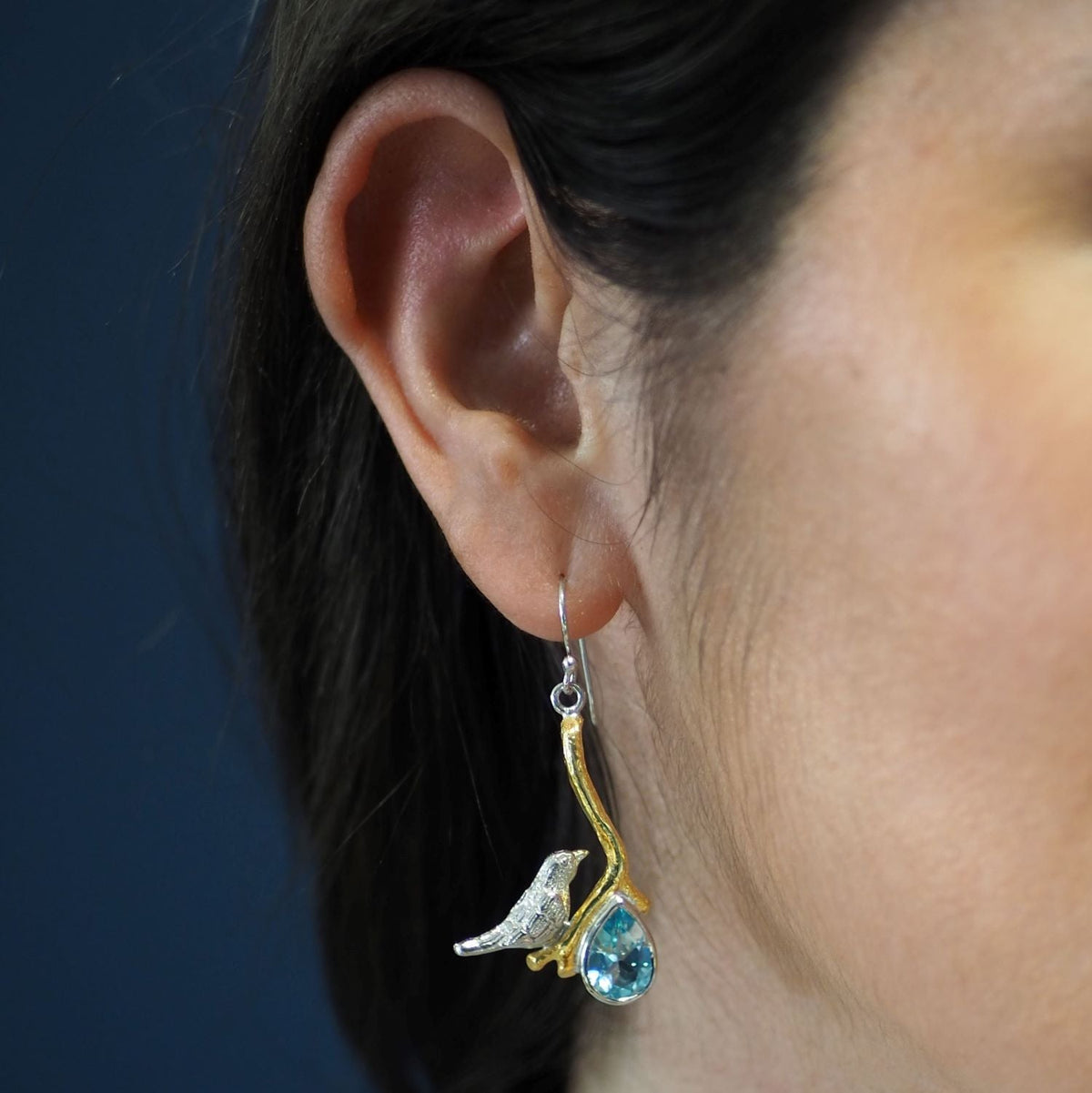 Close-up of a woman's ear wearing unique statement earrings. The earring features a small bird figure perched on a curved, gold-toned branch with a blue teardrop gemstone hanging below. The background is a simple, solid color. She is wearing the Lovebird