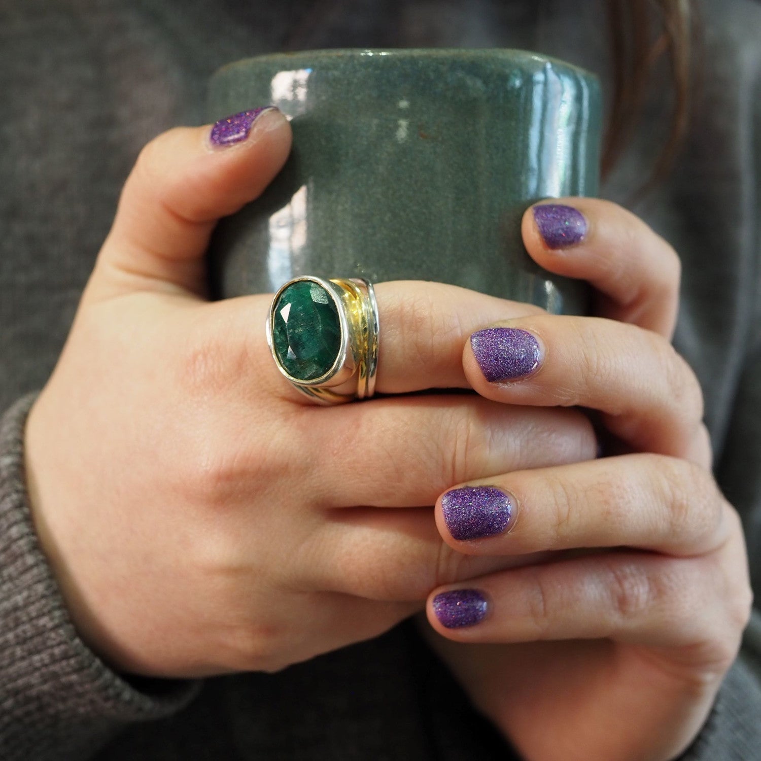 A person with glittery purple nail polish holds a green mug with both hands. They are wearing a large sterling silver & gold Party Ring in various gemstones by Gallardo & Blaine Designs on one hand and another adjustable ring on their middle and ring fing