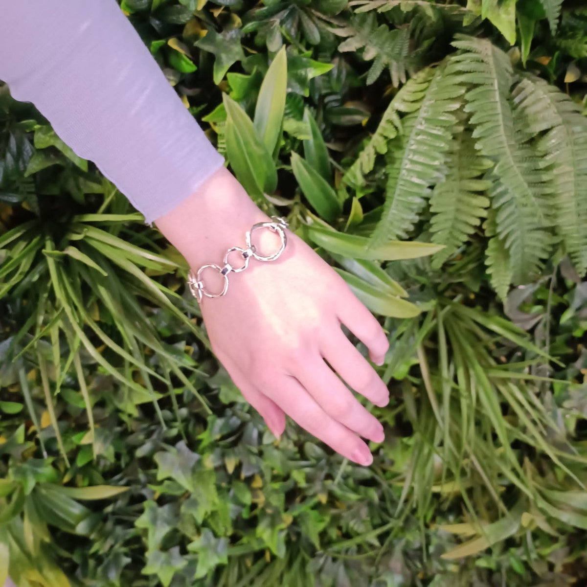 A hand with a Gallardo & Blaine Designs Sea Honey Bracelet Silver featuring thick silver links is extended against a background of lush green foliage. The person is wearing a long-sleeved, light purple top, showcasing one of the new designs.