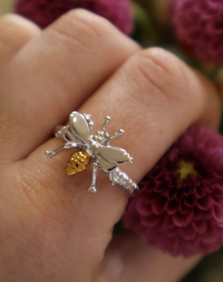 A close-up of a hand wearing the Wildlife Bee Ring Silver & Gold by Watermelon Tropical, featuring gold accents and an adjustable fit, held near vibrant purple flowers.
