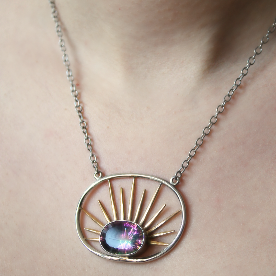 A close-up of a person's neck wearing Gallardo & Blaine Designs' Goddess Necklace Large in Various Gemstones, featuring a silver chain and sun-inspired pendant with a purple gem and gold vermeil rays—a striking statement piece.